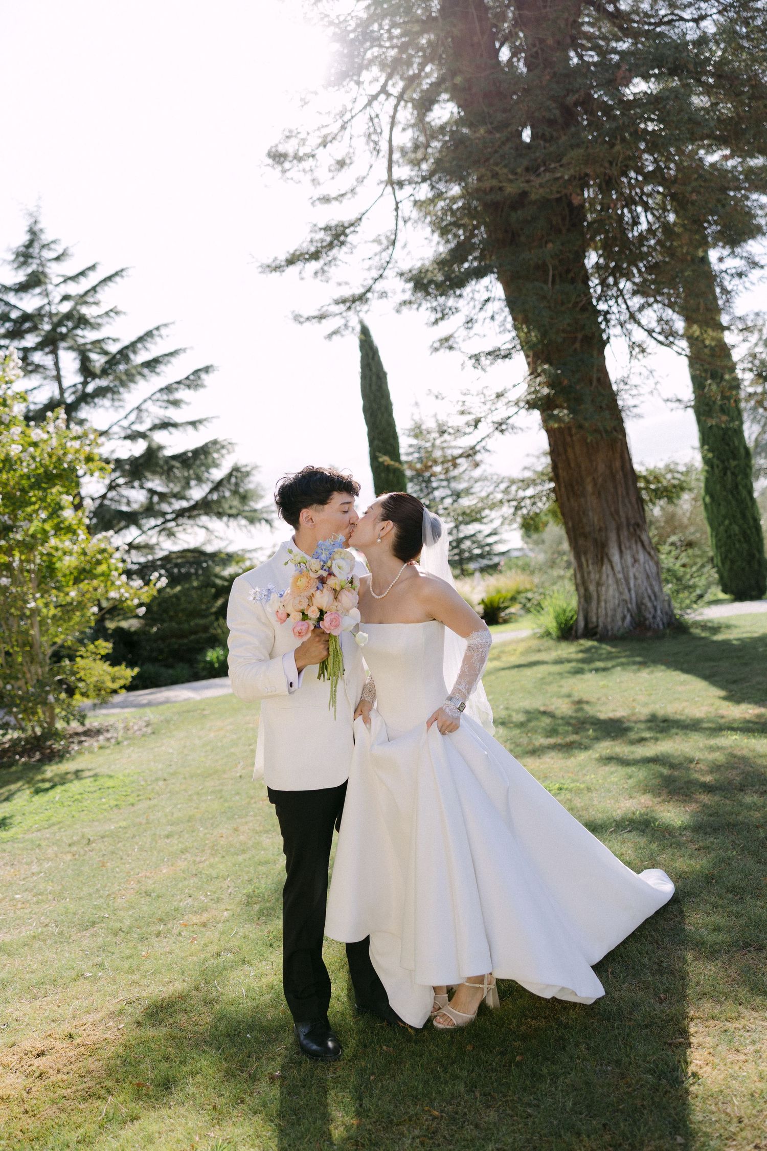 Bride and groom share a romantic kiss outdoors on a sunny day, bride holding a pastel bouquet in a white gown.