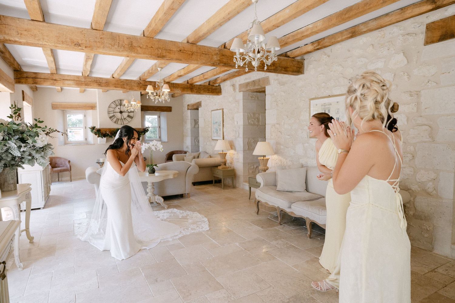 Two brides in white gowns preparing in a rustic French venue with exposed beams and stone walls.