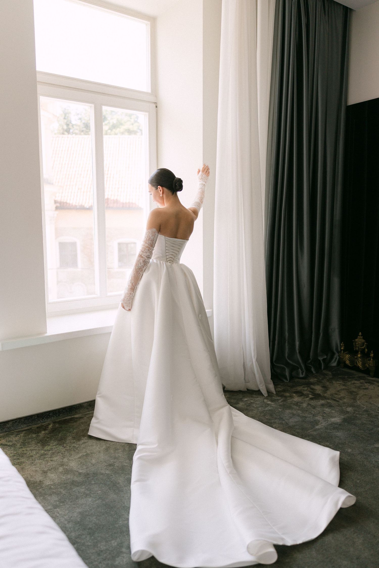 Elegant bride in a strapless white ball gown with lace gloves standing by a large window.