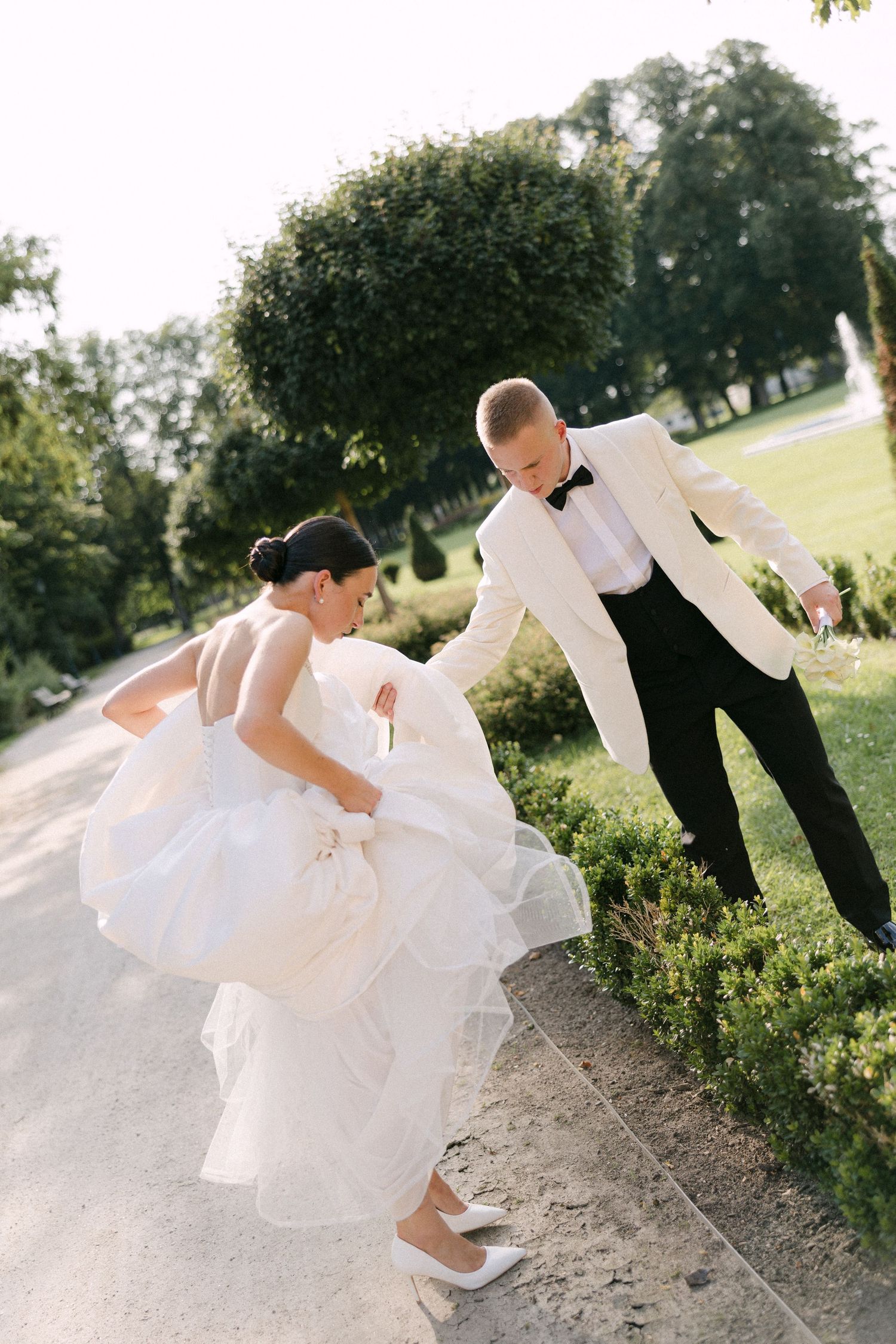 Bride lifting her white ruffled gown while groom in white tuxedo assists her on a garden pathway.