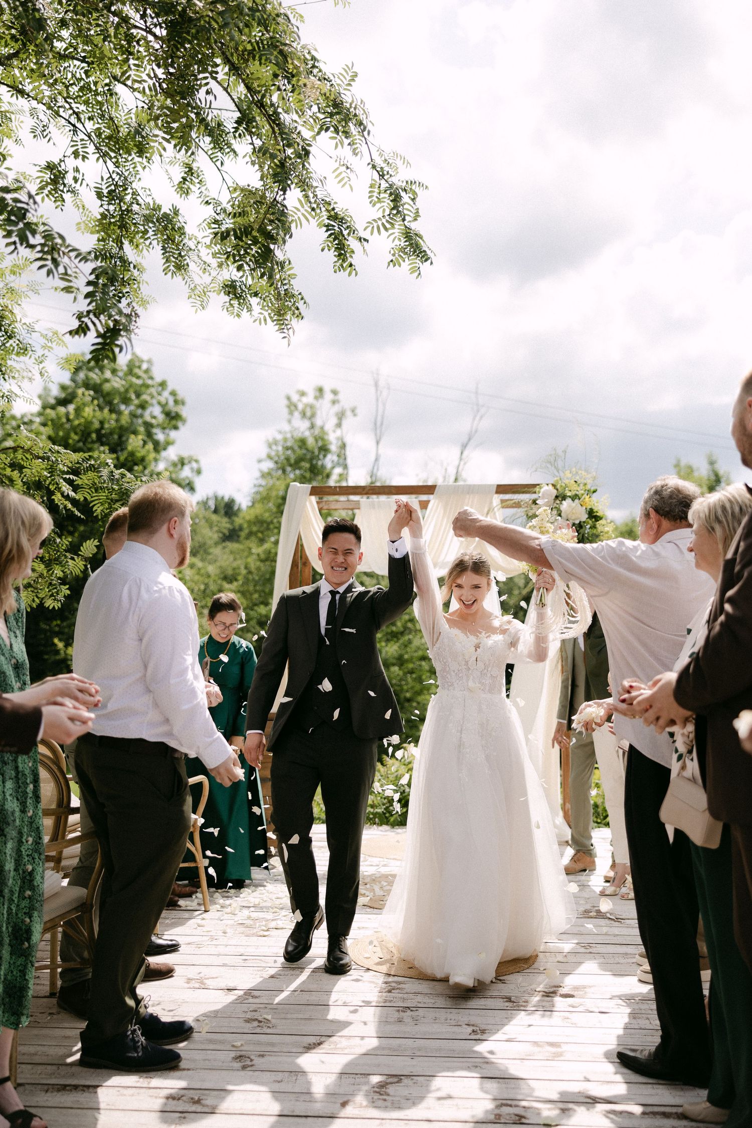 Newlyweds celebrating their recessional with raised hands as guests cheer at an outdoor wood deck ceremony.