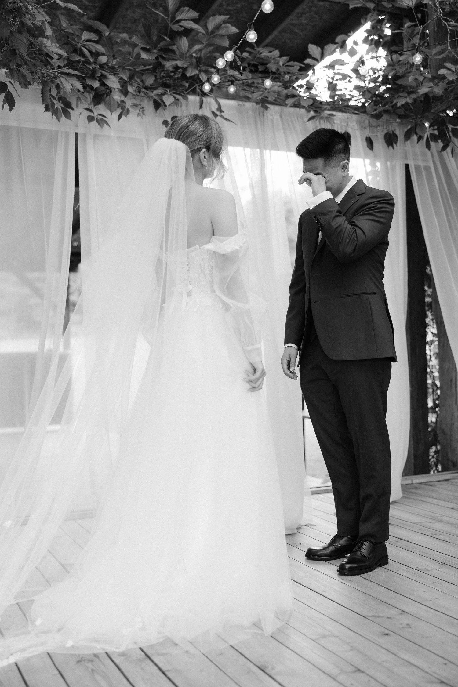 Black and white photo of a groom wiping tears as bride stands before him at wedding ceremony altar.