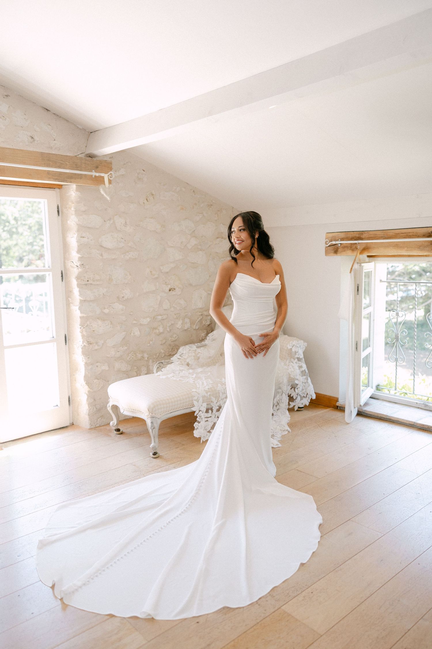 Elegant bride in strapless fitted wedding gown with long train standing in sunlit stone room.