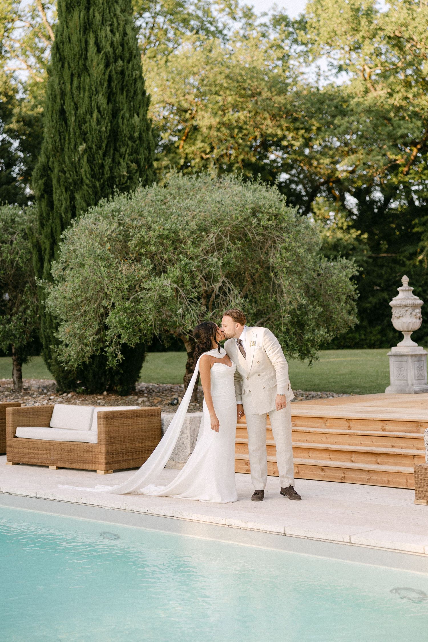 Bride and groom share a romantic kiss poolside at an elegant outdoor wedding venue with lush greenery.