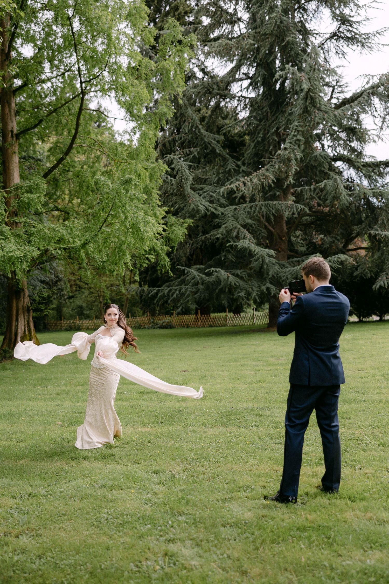 Bride in elegant gown poses outdoors while groom photographs her in lush green park setting.