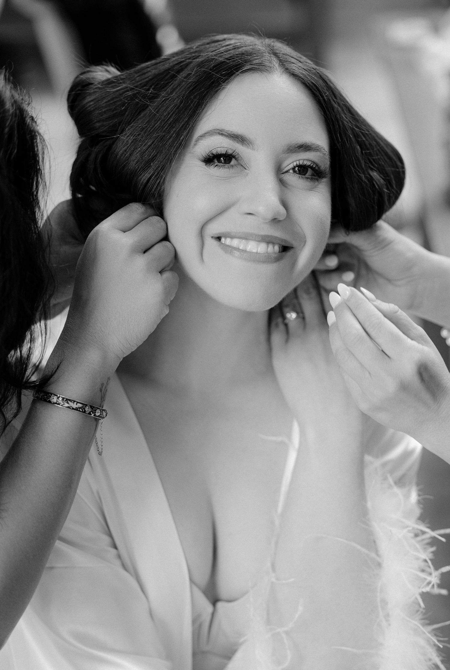 Black and white portrait of smiling bride having earrings put on during wedding preparation.