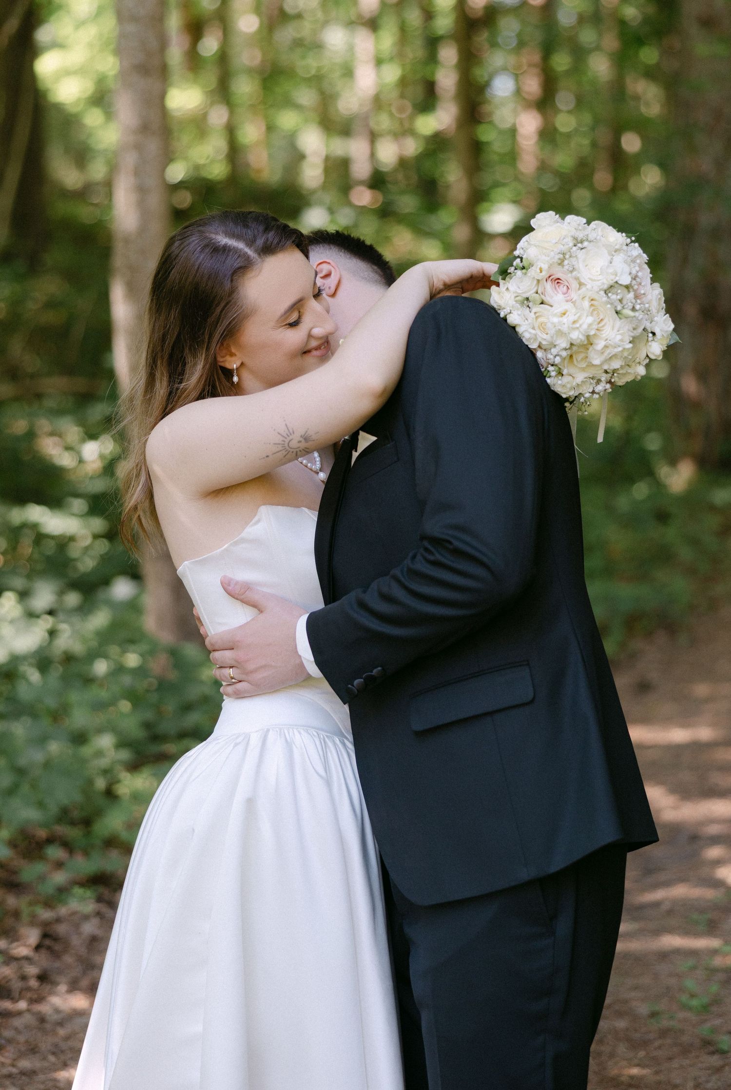 Bride and groom share tender kiss embrace in forest holding white floral bouquet on wedding day.