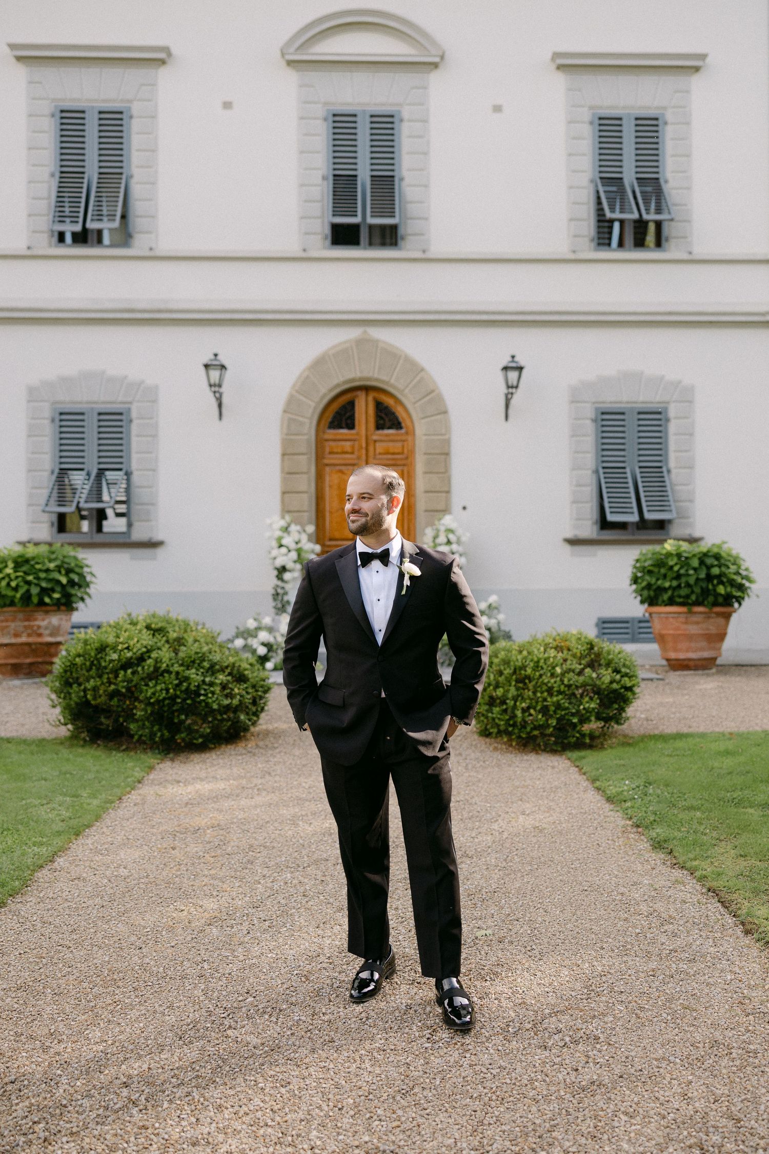 Groom in black tuxedo with bow tie stands confidently on gravel path before elegant white Italian villa.