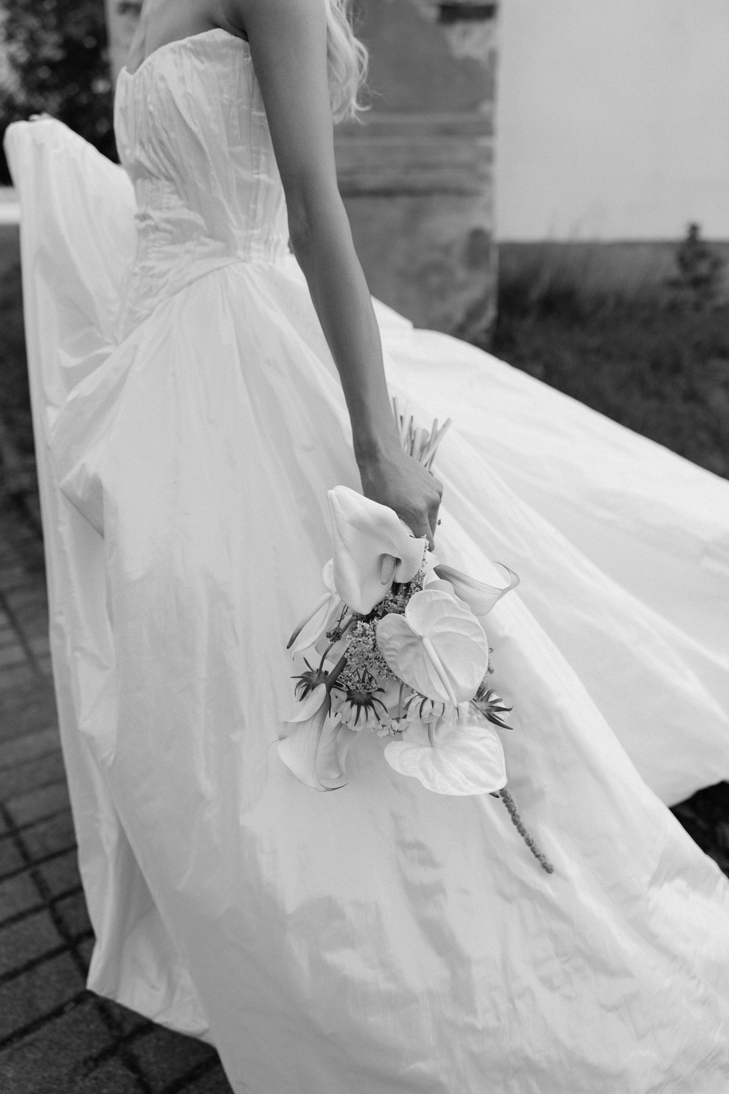 Black and white photo of a bride in a flowing strapless gown holding a white floral bouquet outdoors.