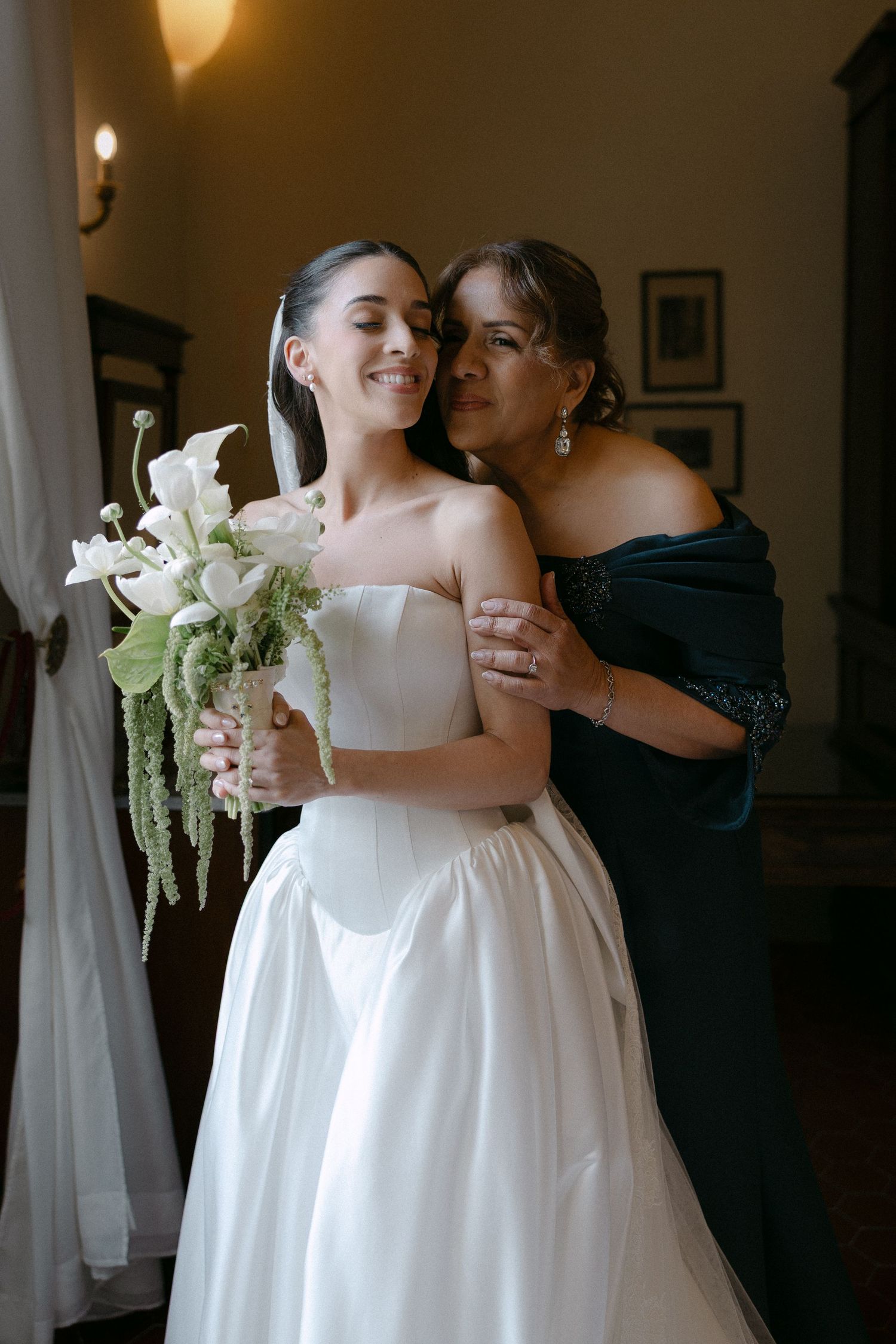 Bride in strapless white gown holding calla lily bouquet embraced by mother in teal dress indoors.