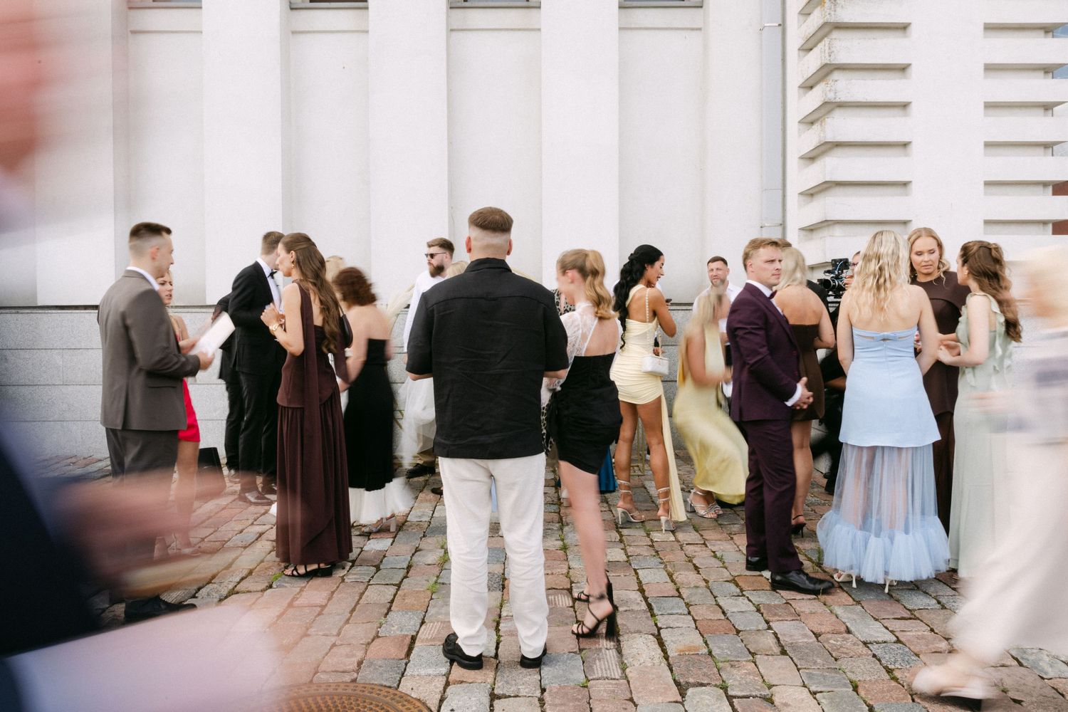 Elegantly dressed wedding guests mingling outside white classical building on cobblestone plaza.