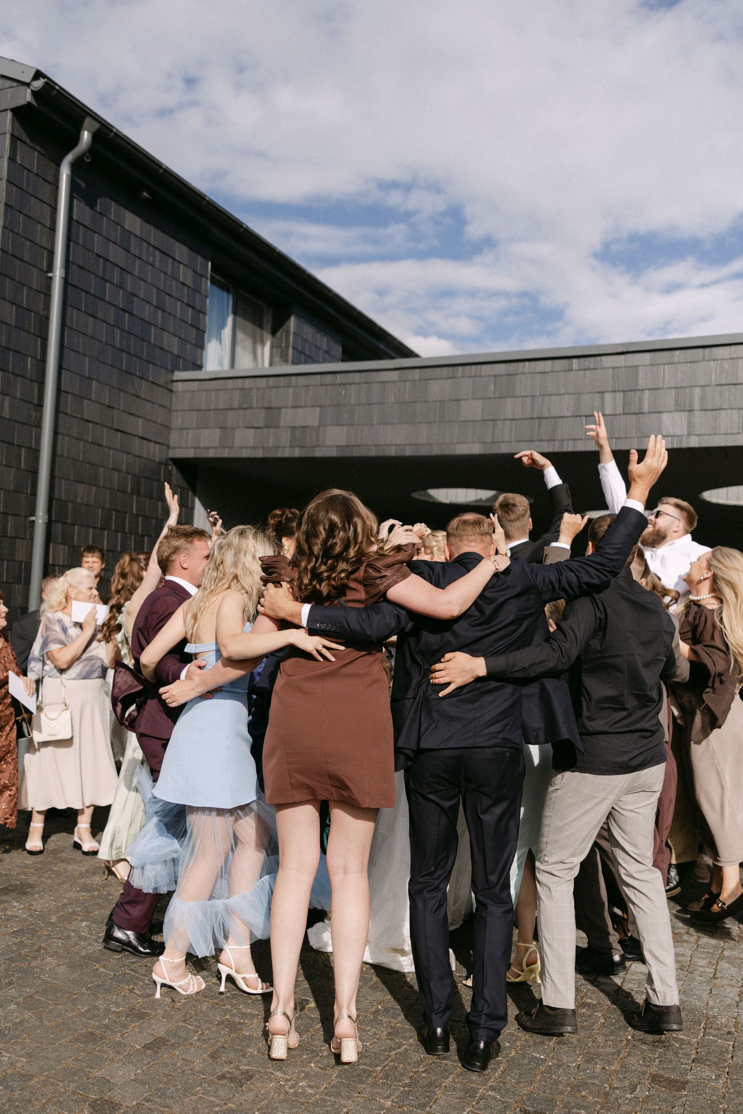 Wedding guests hugging and celebrating together outside a modern dark building under blue sky.