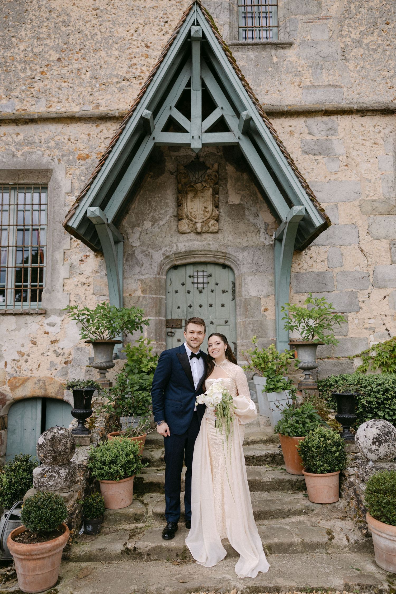 Bride and groom pose on stone steps before a historic chateau entrance with ornate wooden canopy.