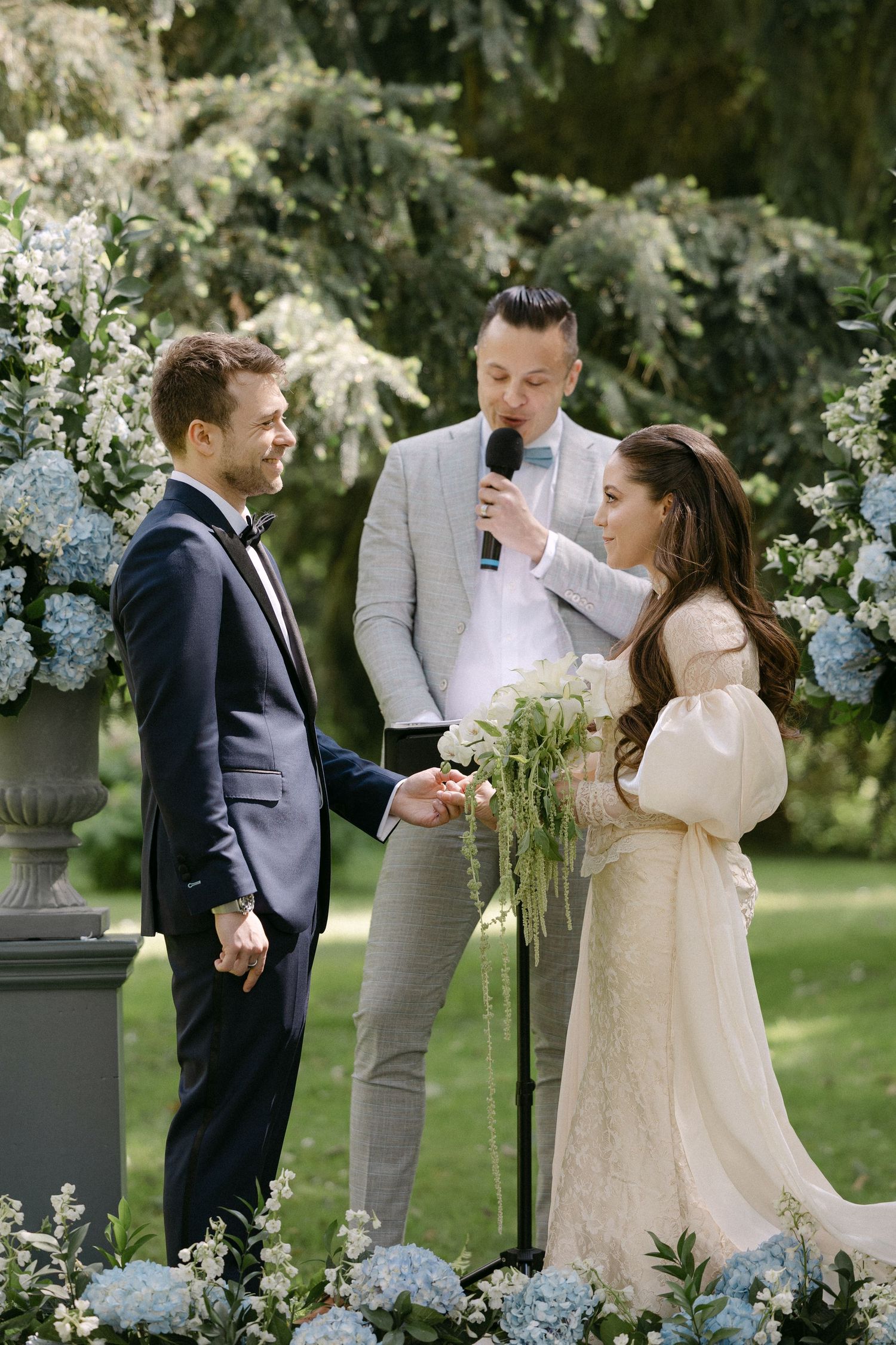 Bride and groom exchange vows outdoors with officiant, surrounded by blue hydrangea floral arrangements.