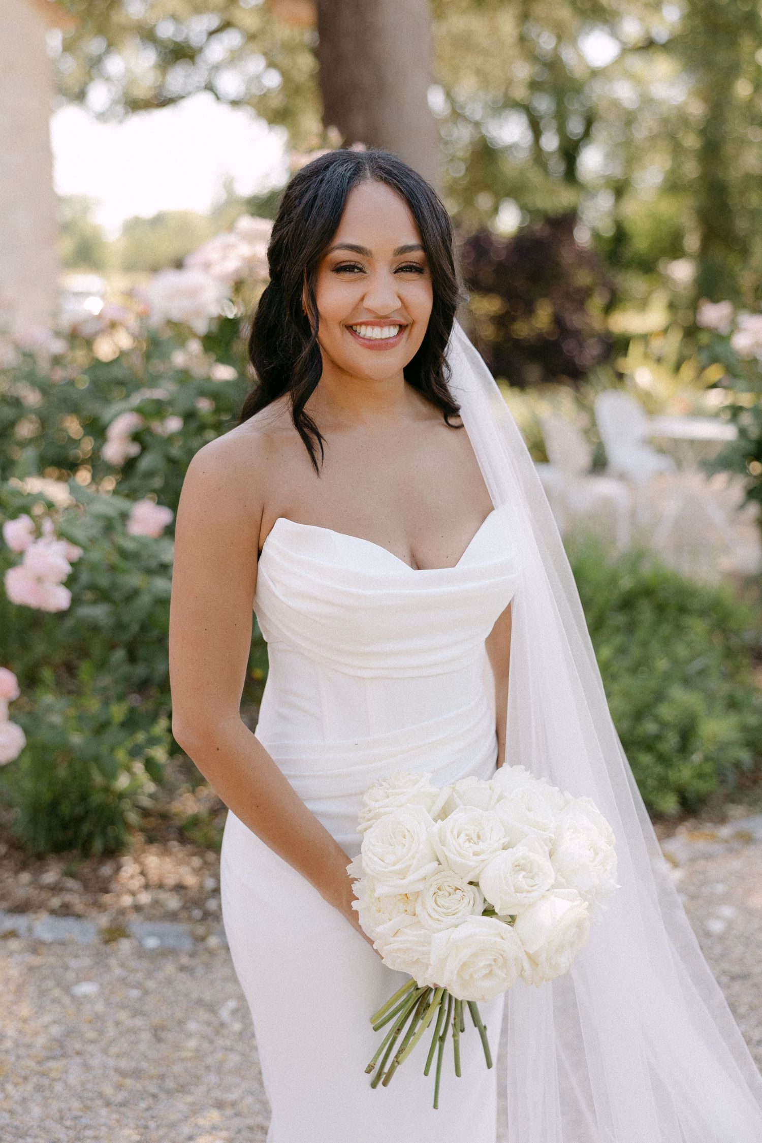 Smiling bride in strapless white gown holds all-white rose bouquet in garden setting.