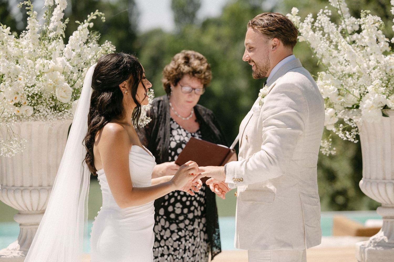 Bride and groom exchange rings during outdoor wedding ceremony with white floral arrangements and officiant.