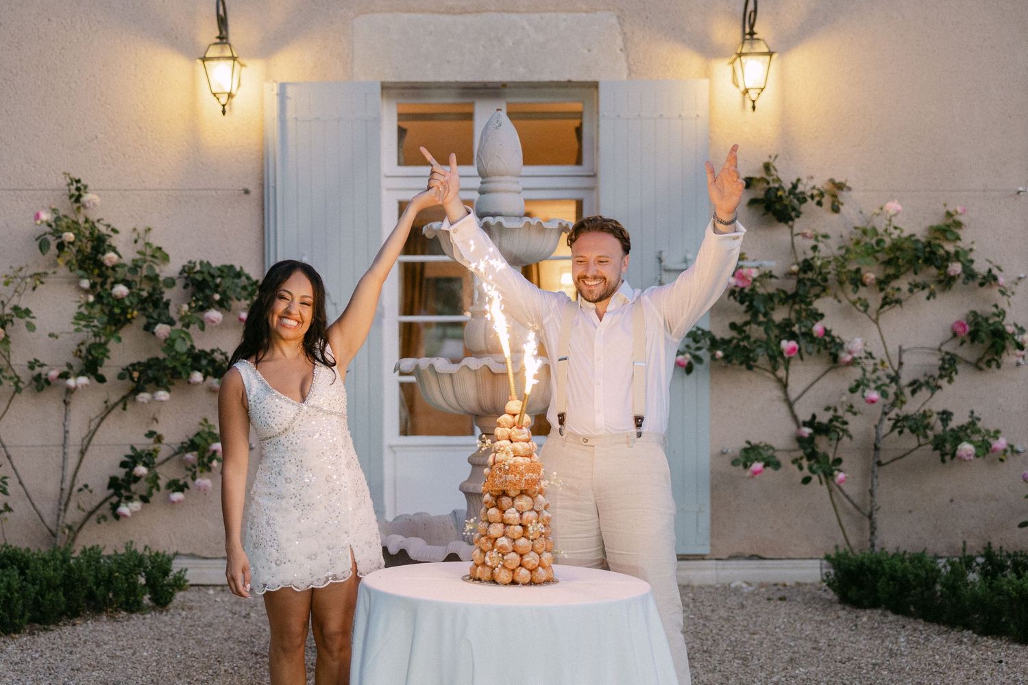 Bride and groom celebrating with sparklers beside a French croquembouche wedding cake at dusk.