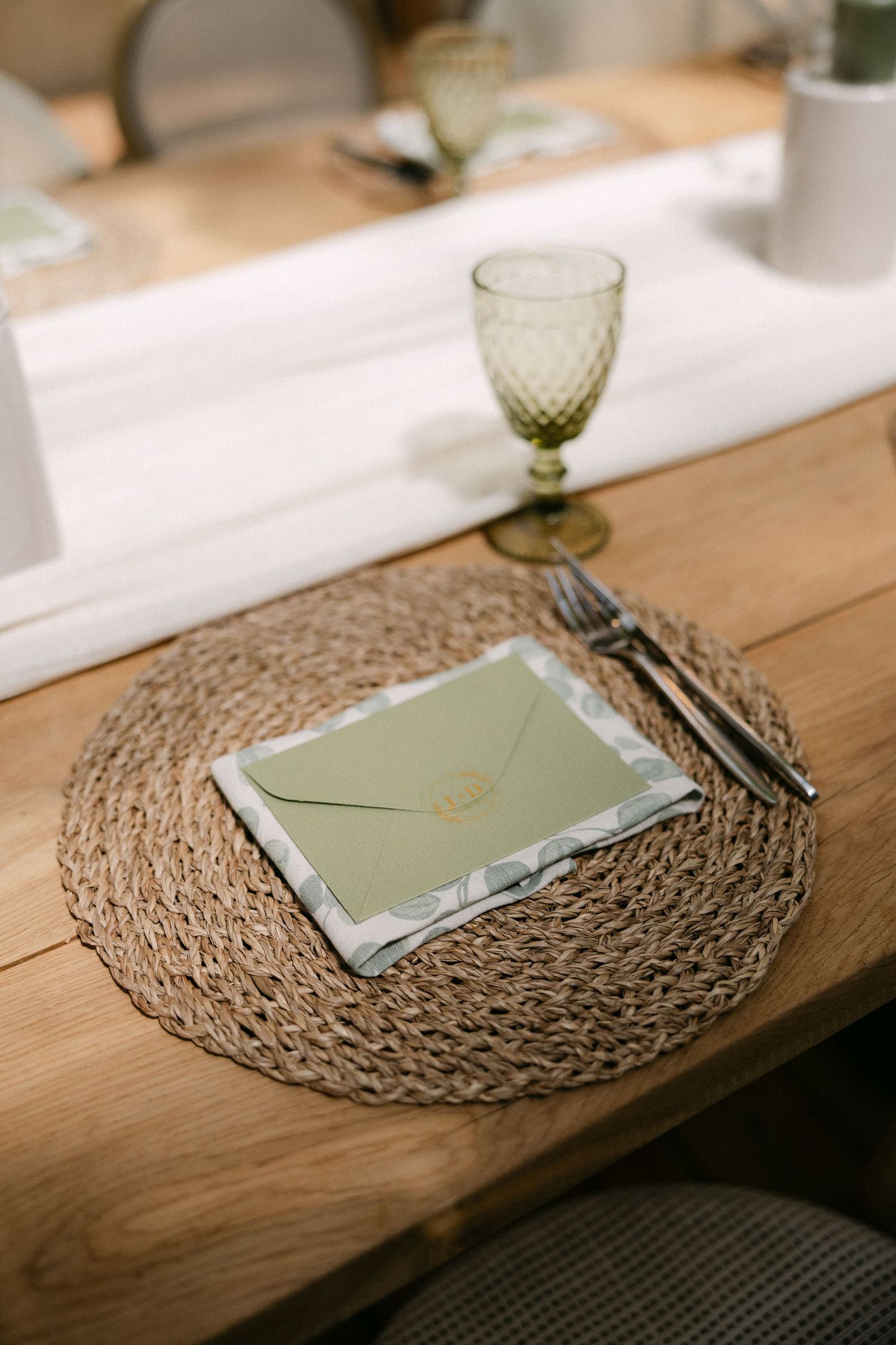 Elegant wedding place setting with sage green envelope on woven placemat, silverware, and green goblet on wood table.