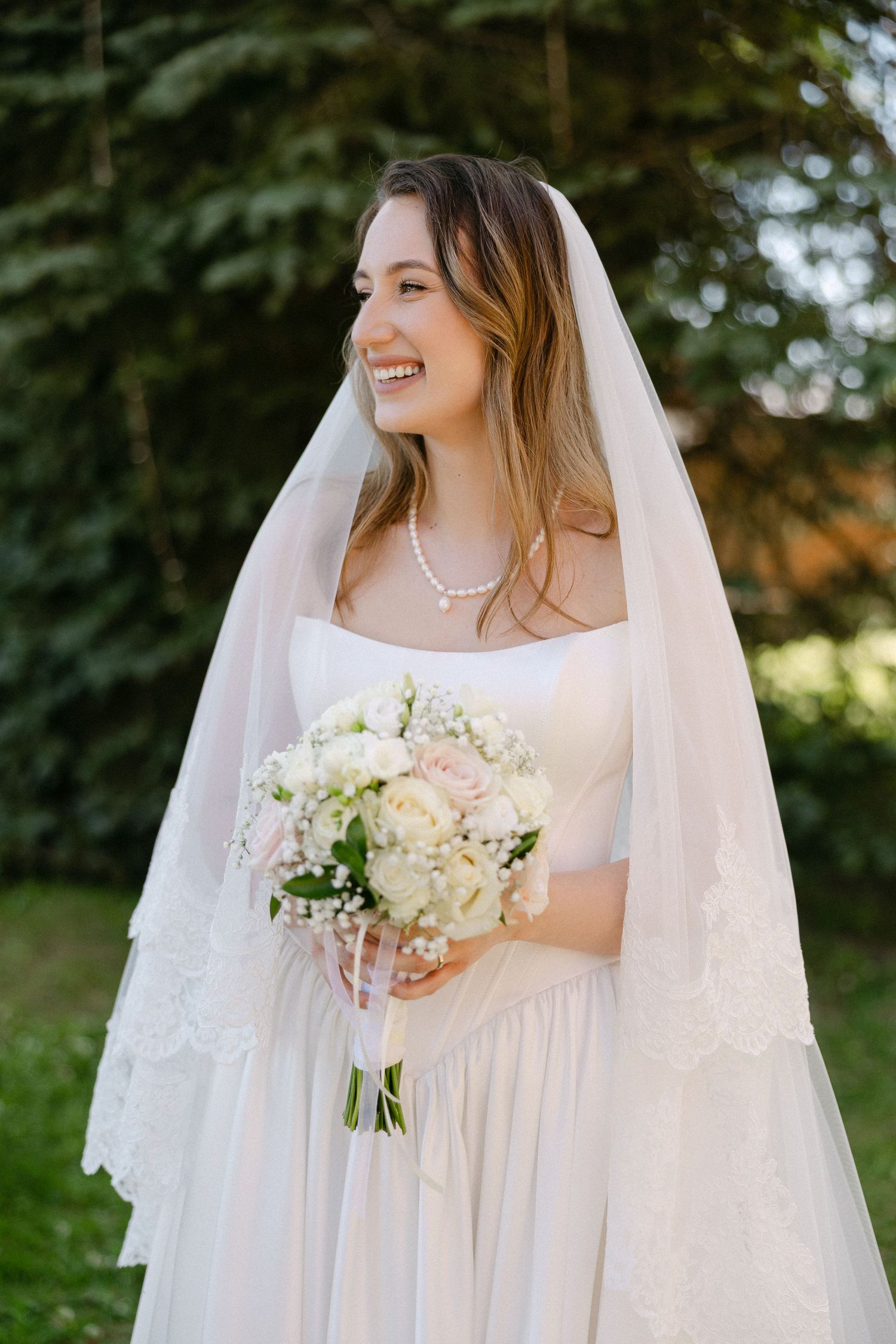 Smiling bride in white gown and veil holding elegant bouquet of roses outdoors.