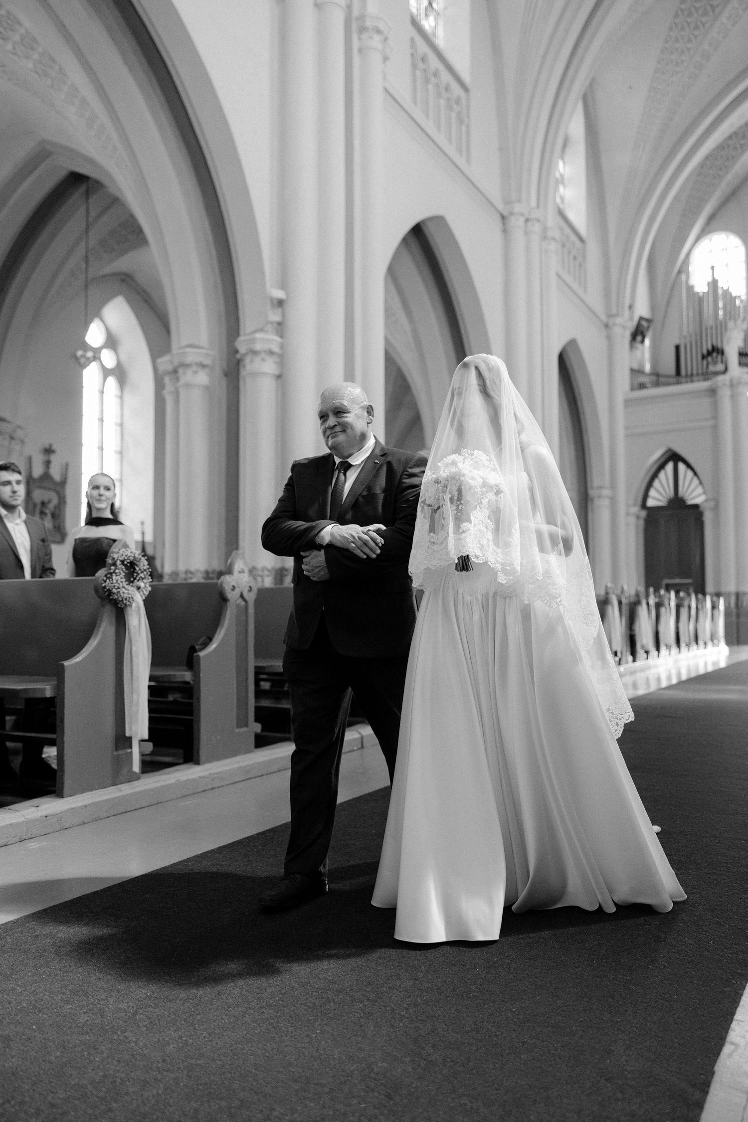 Black and white photo of bride in flowing white gown and veil walking down church aisle with father.