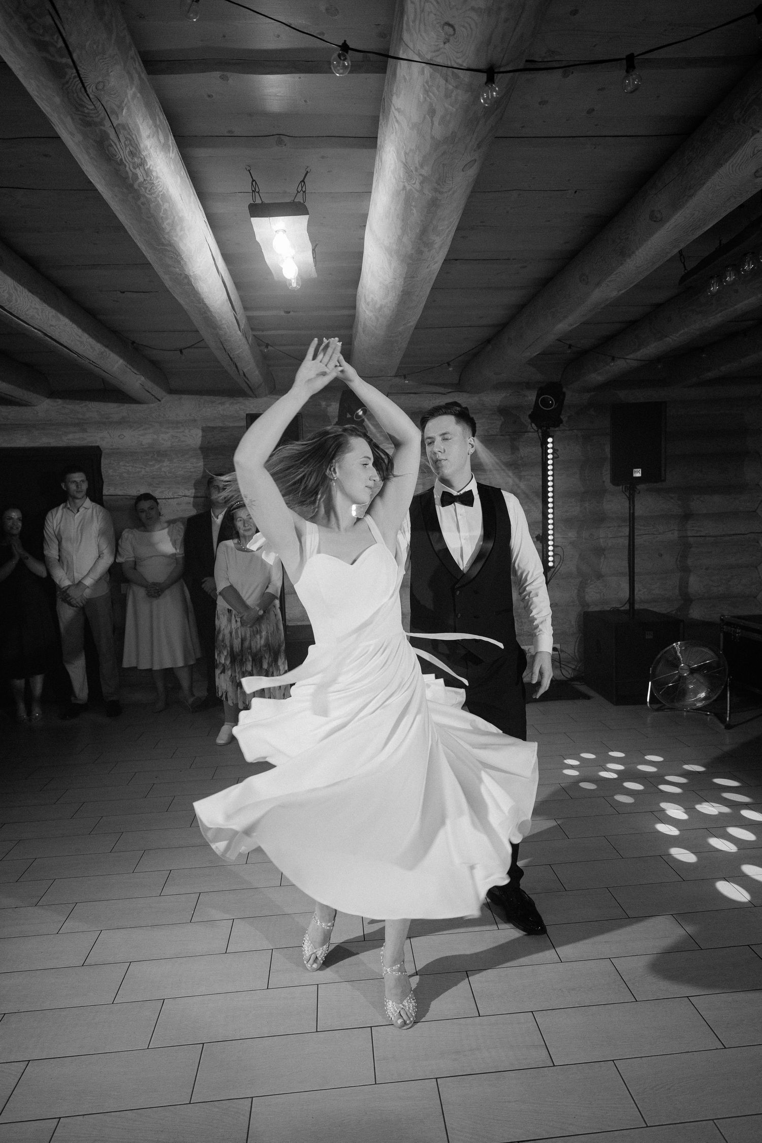 Bride and groom share first dance in black and white photo, bride twirling in flowing white dress under rustic wooden ceiling.