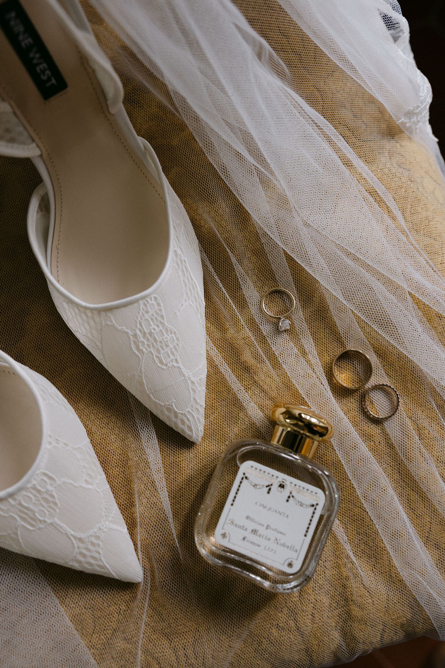 Nine West white lace wedding shoes with perfume bottle and gold rings on veil-draped surface.