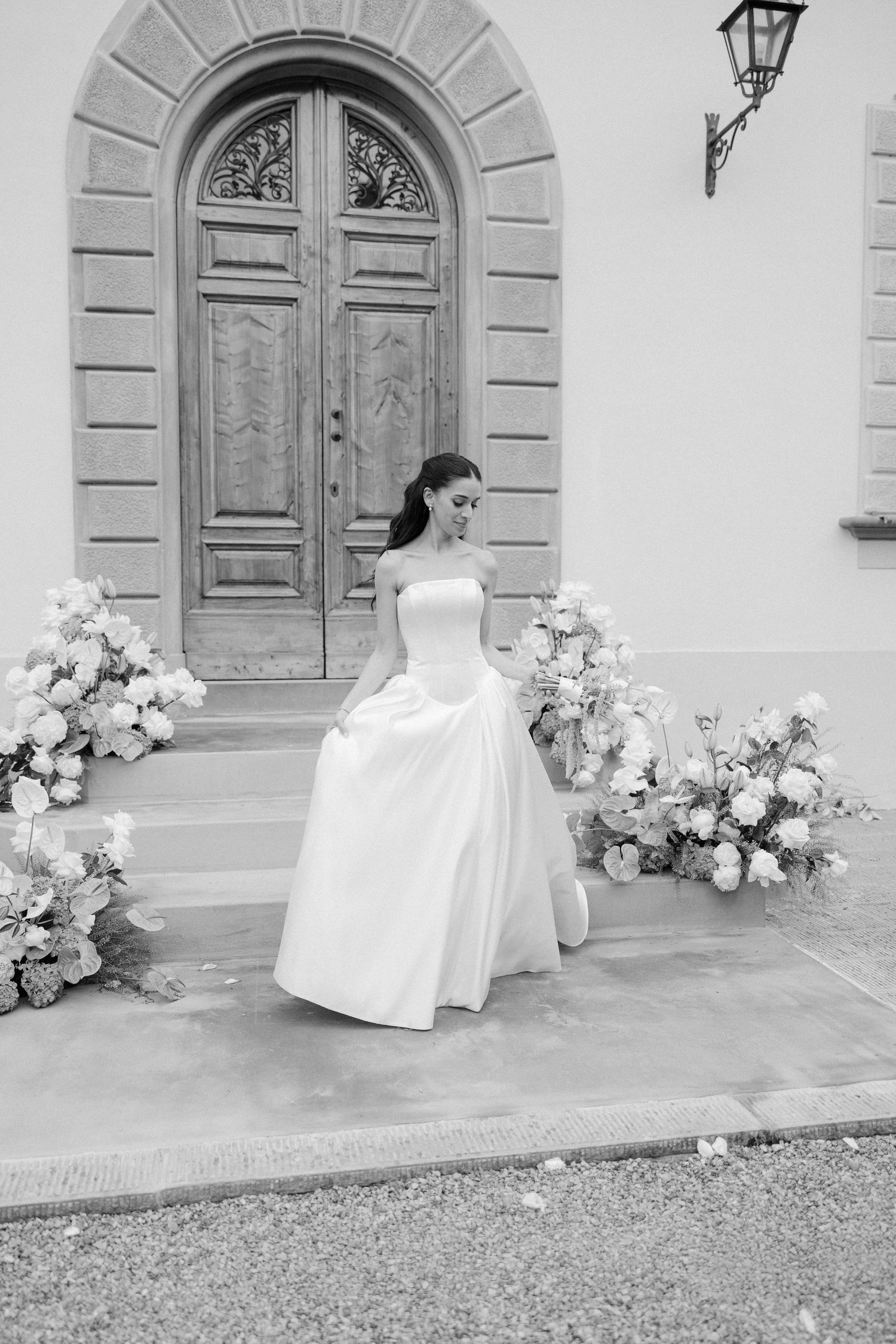 Black and white photo of bride in strapless gown on steps flanked by floral arrangements at villa.