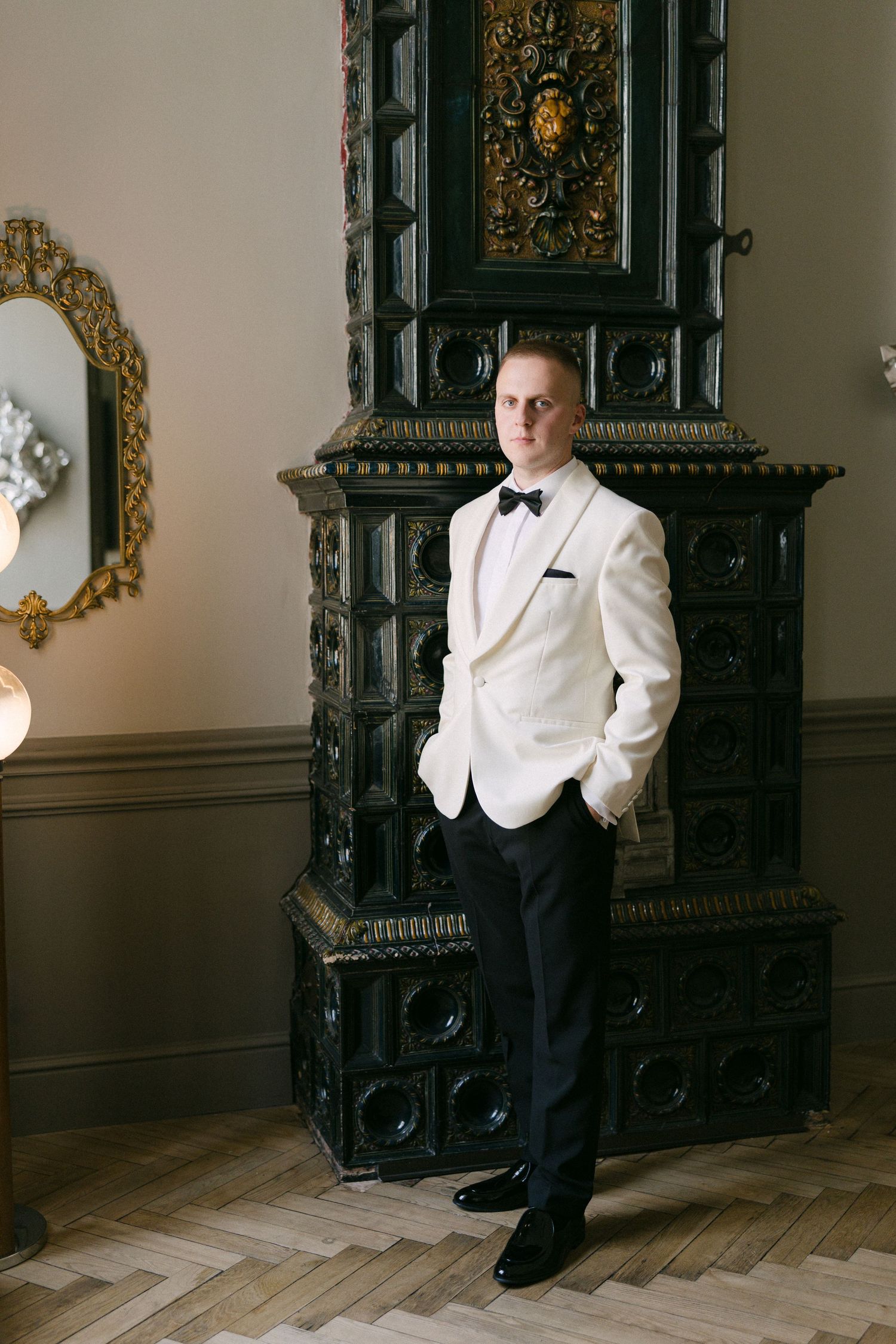 Groom in white tuxedo jacket and black bow tie stands before ornate dark antique fireplace on herringbone floor.