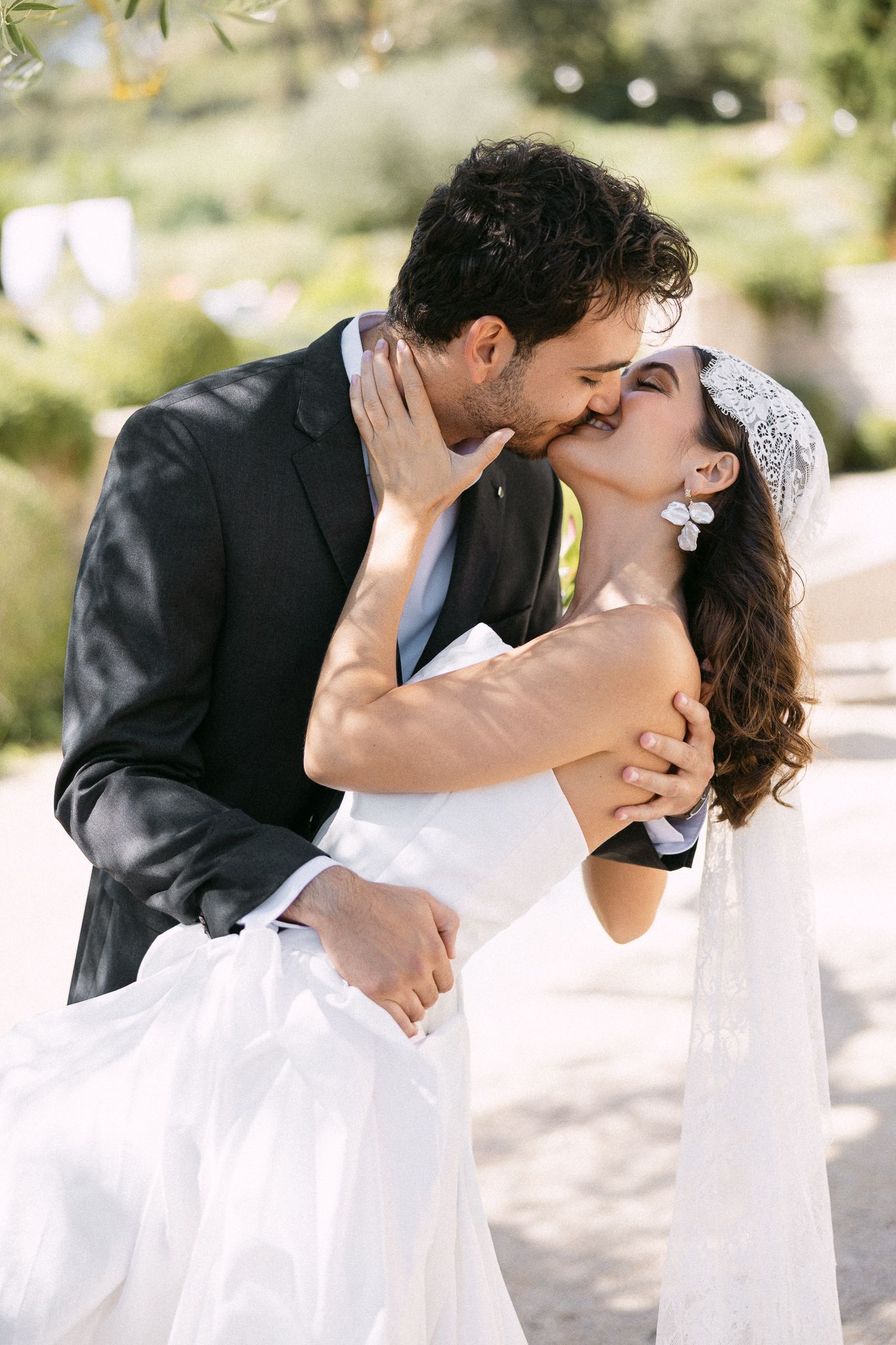 A bride and groom share a romantic kiss outdoors, with the bride in a white gown and veil embracing her suited groom.