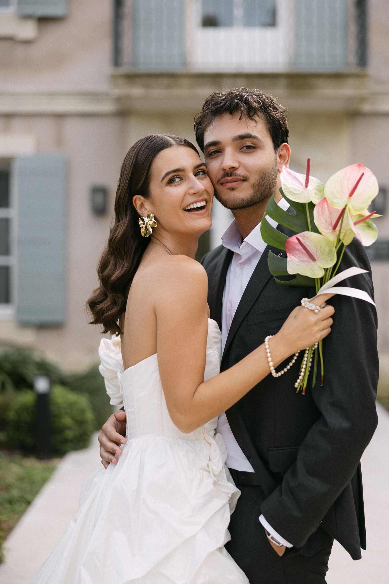 Joyful bride and groom embracing while holding anthurium bouquet outside French estate.