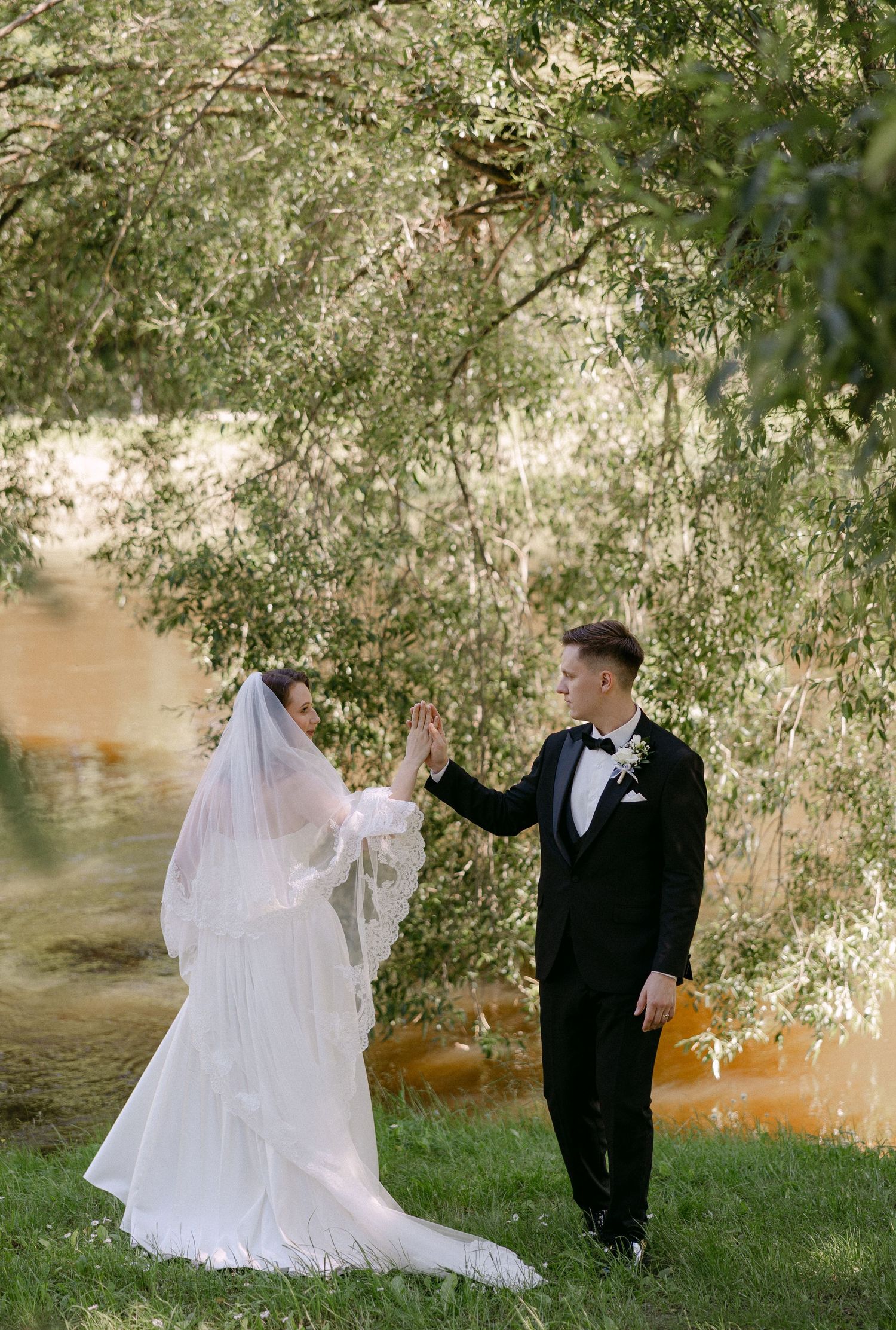 Bride and groom hold hands by willow tree near river in elegant outdoor wedding portrait.