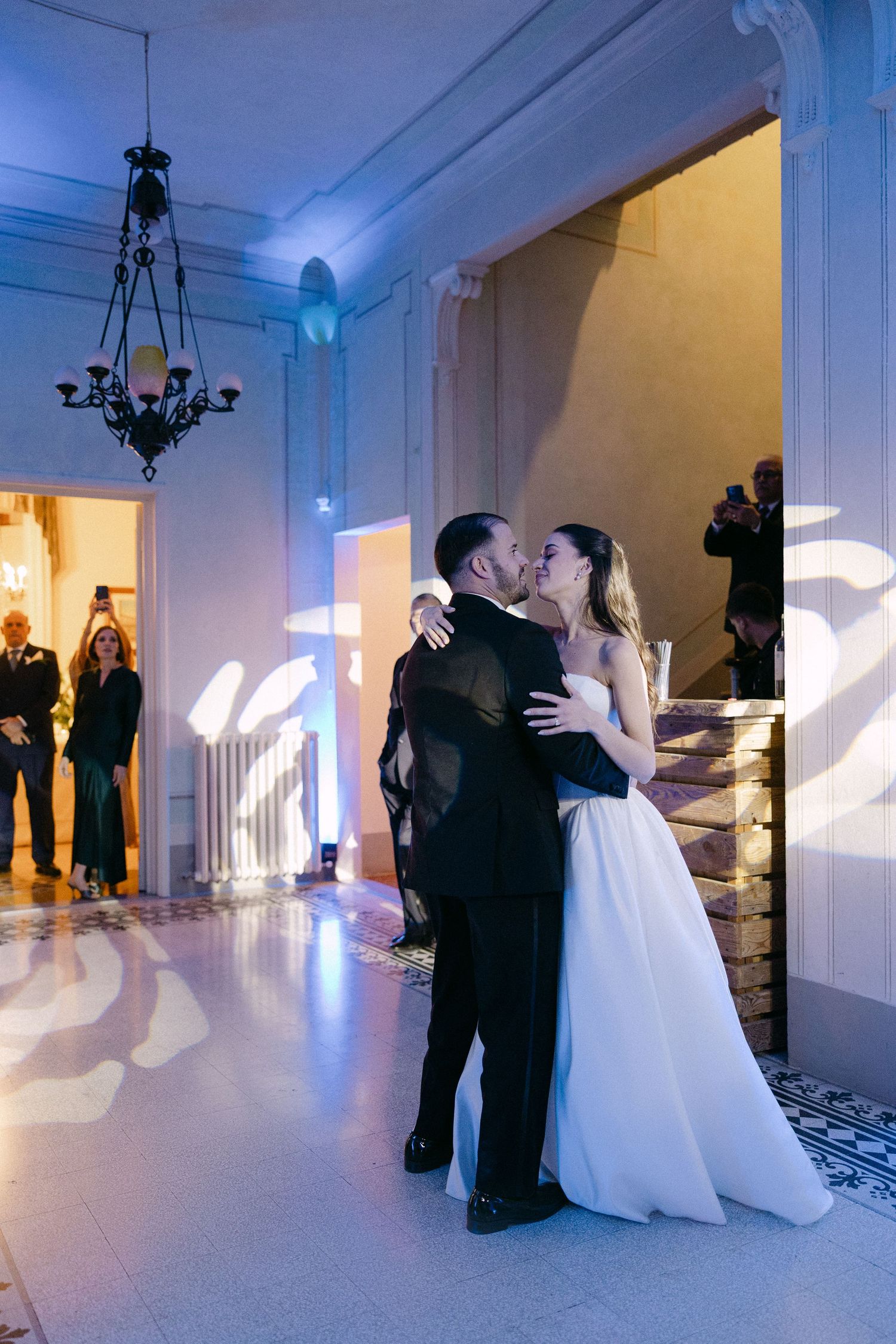 Bride and groom share romantic first dance kiss in ornate ballroom with blue ambient lighting.