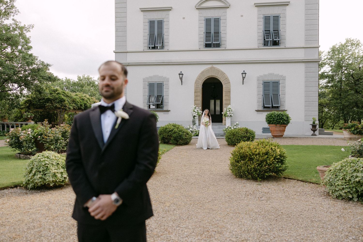 Groom in black tuxedo stands in foreground as bride approaches elegant Italian villa during outdoor wedding ceremony.