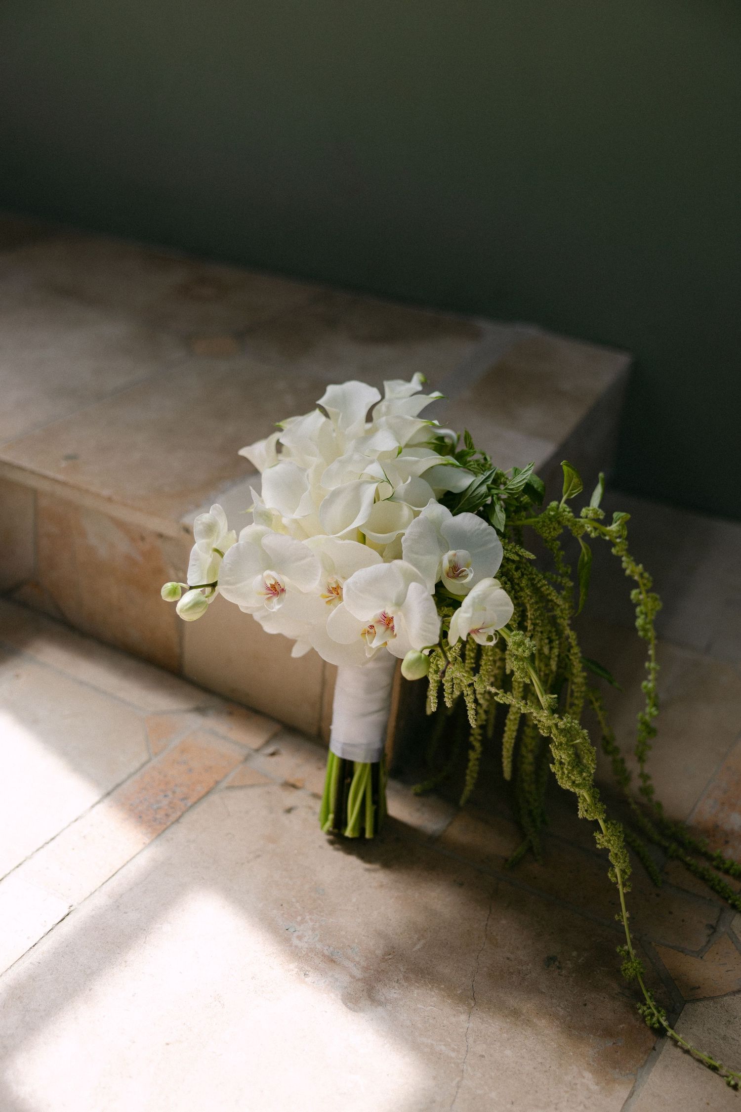 Elegant bridal bouquet of white orchids and calla lilies with trailing greenery on marble steps.