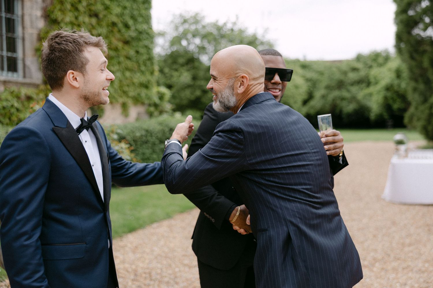 Wedding guests in formal suits greeting each other with hugs outside a historic stone venue.