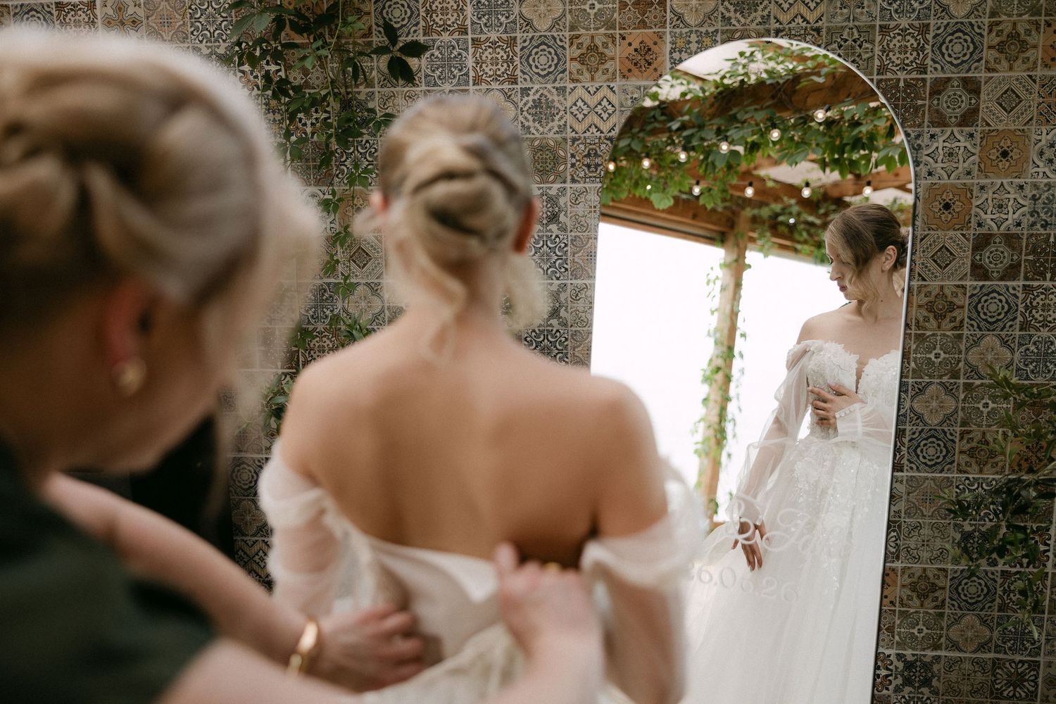 Bride getting her wedding dress fastened by a bridesmaid in front of an ornate tiled mirror.