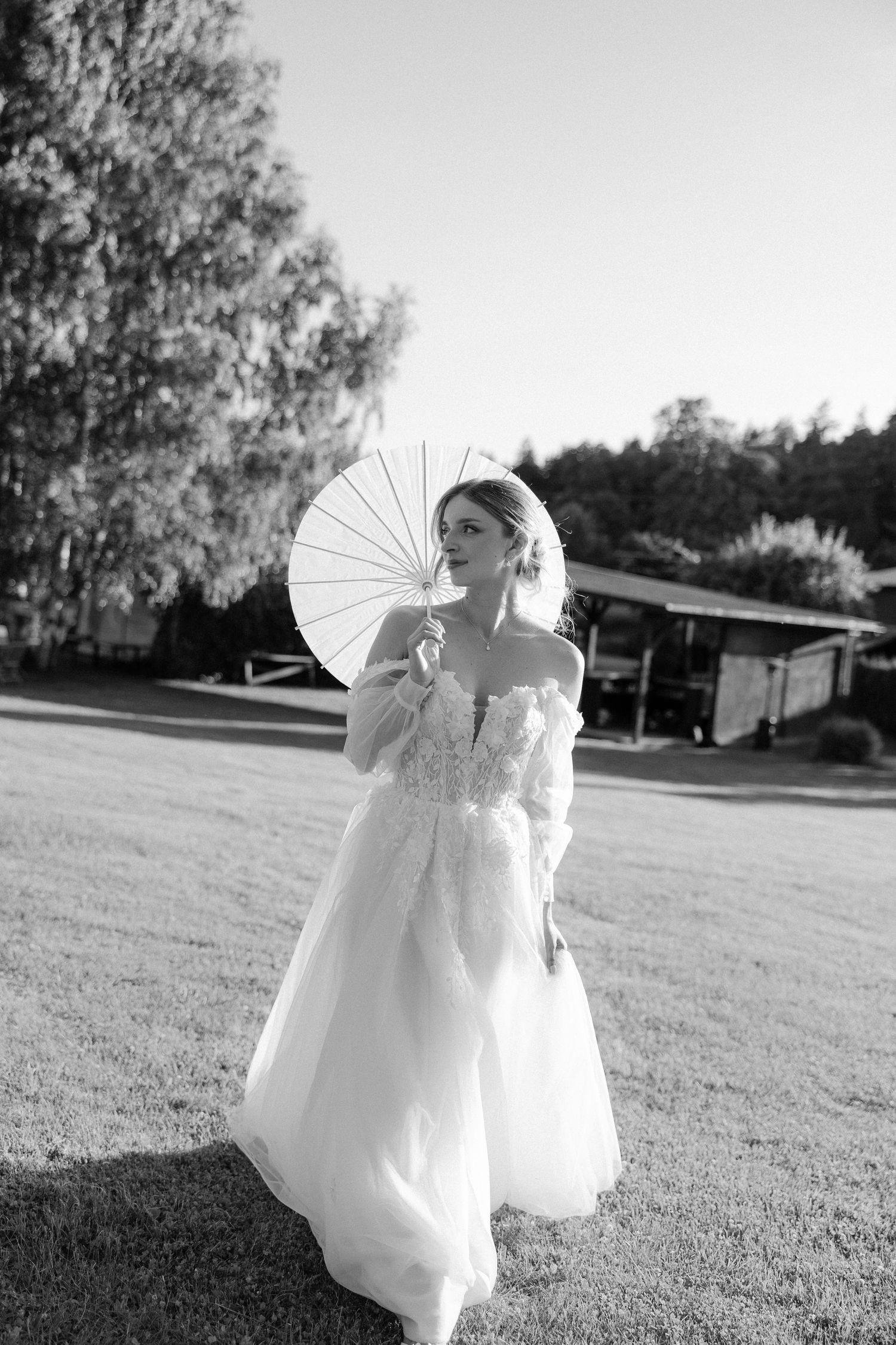 Bride in white tulle gown holds parasol umbrella while standing on green lawn in black and white photo.