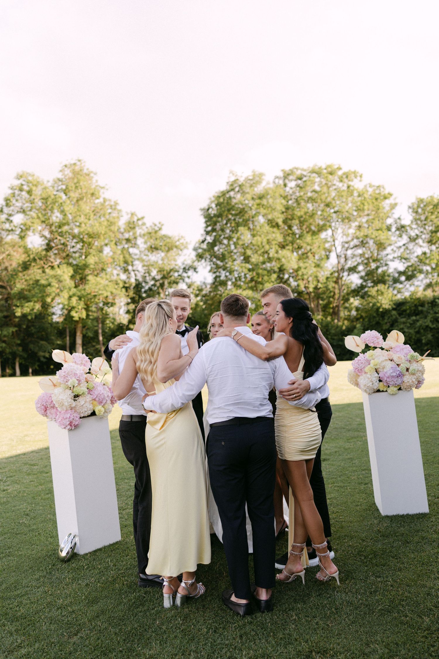 Wedding guests sharing a group hug outdoors on a green lawn with floral arrangements nearby.