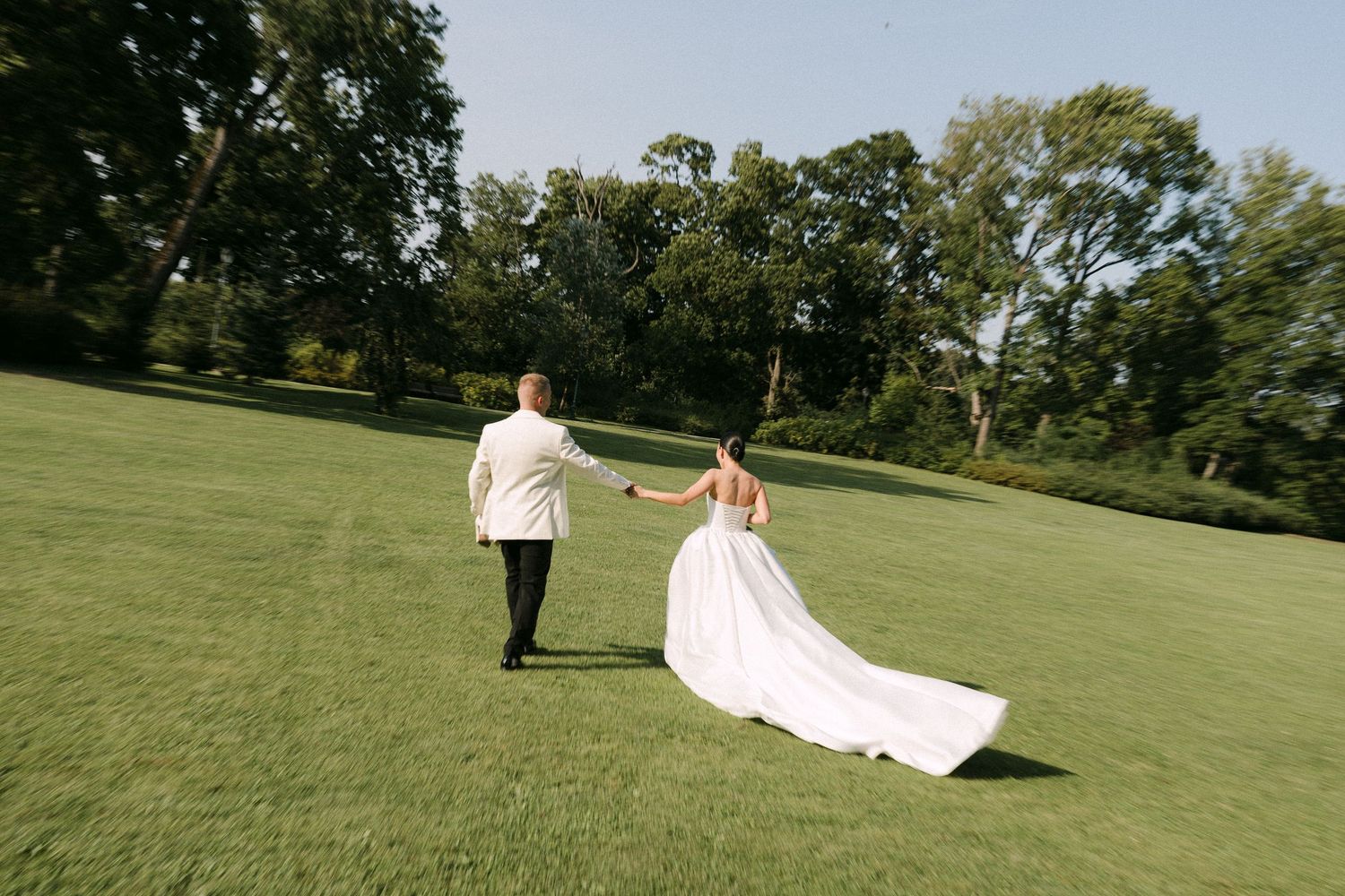 Bride and groom hold hands walking across a lush green lawn surrounded by trees on their wedding day.