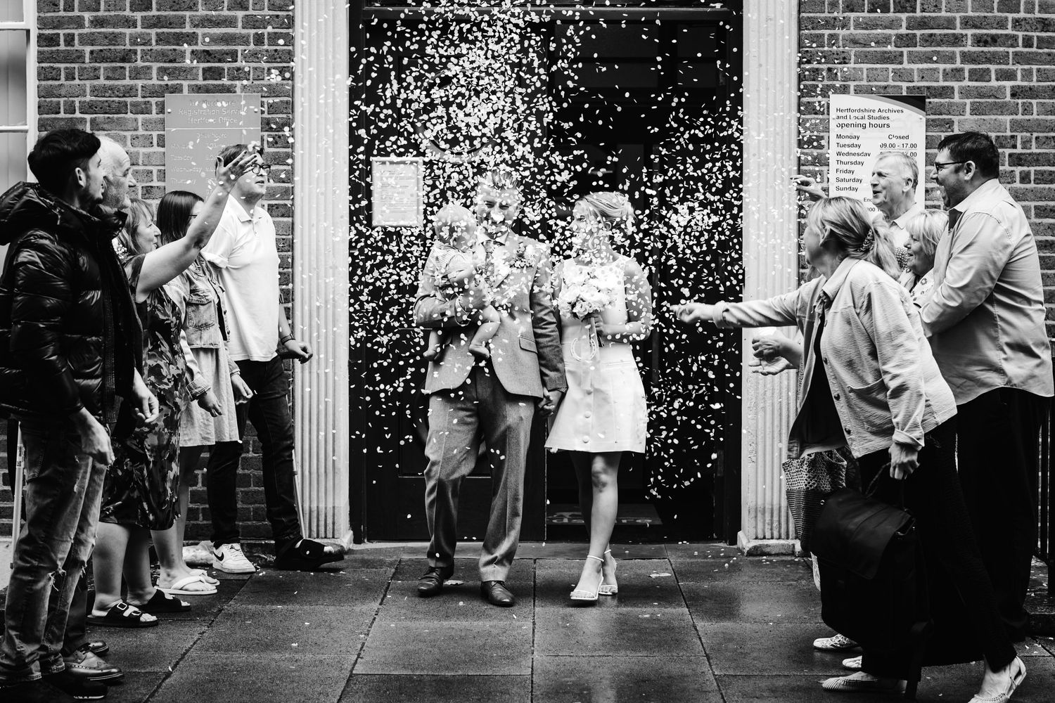 A black and white photo of wedding guests throwing confetti at a brick building entrance.