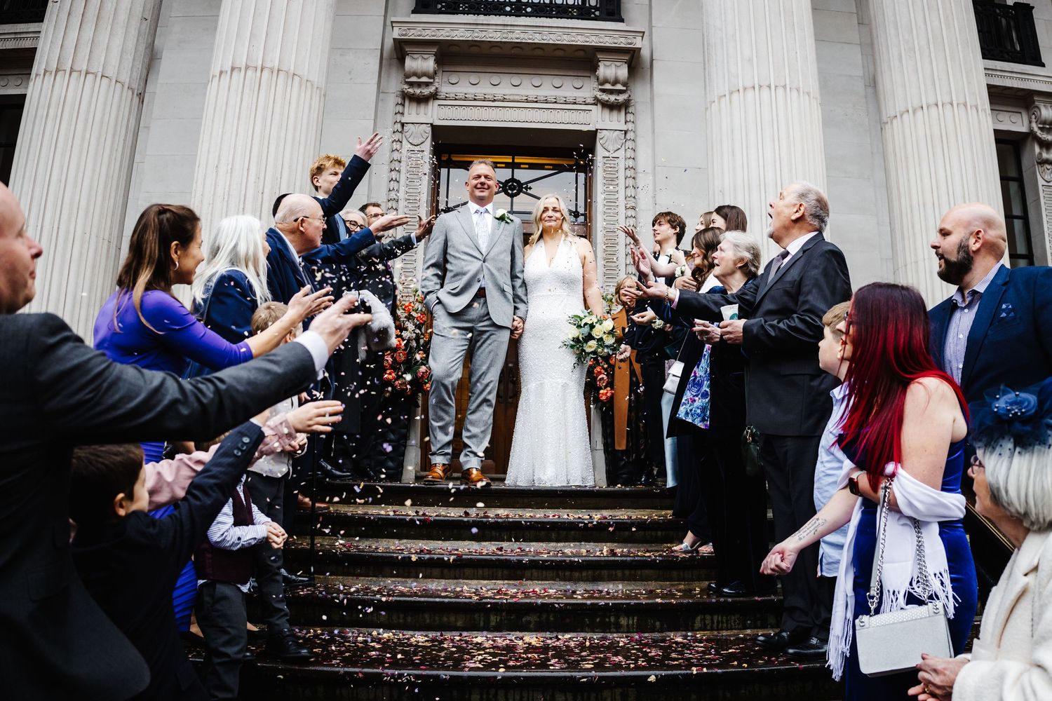 Wedding party and guests celebrate on the steps of a grand building with white columns during a joyful ceremony.