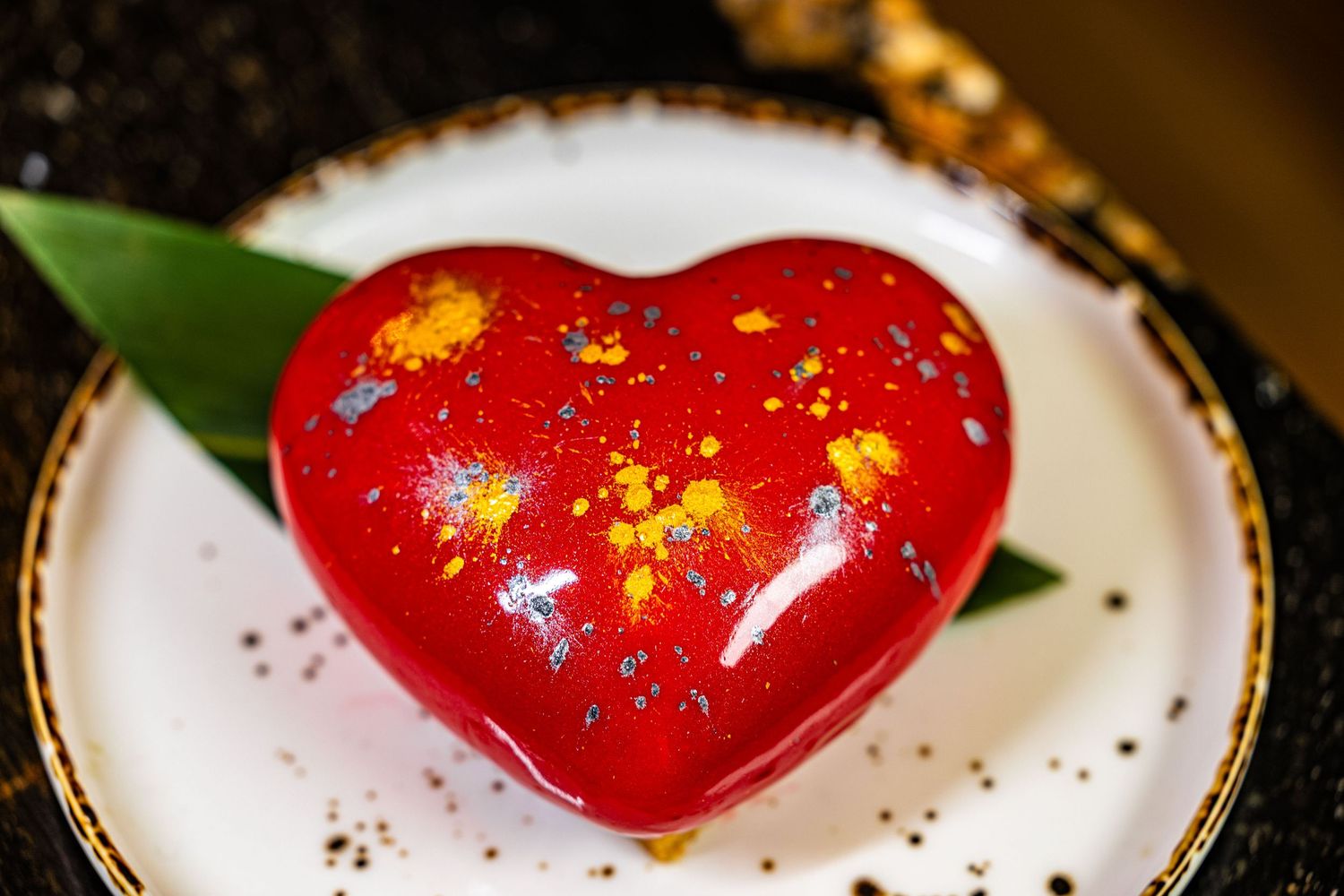 A heart-shaped chocolate dessert with red glaze and gold flakes sits elegantly on a white plate.