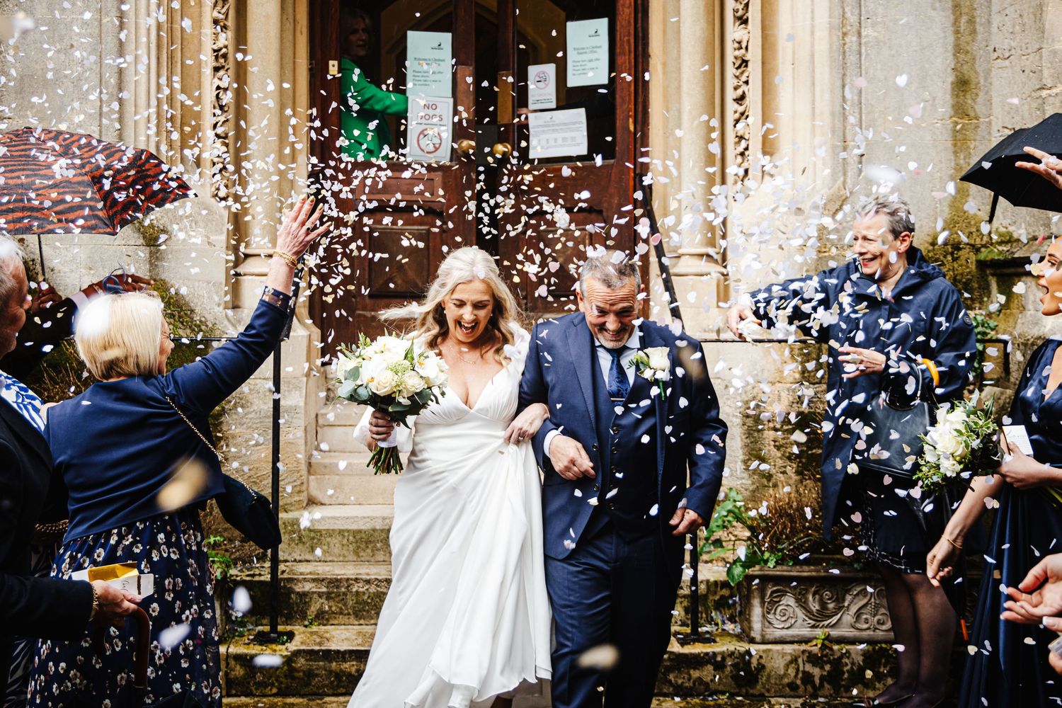 Joyful wedding celebration with guests throwing confetti outside historic stone building.
