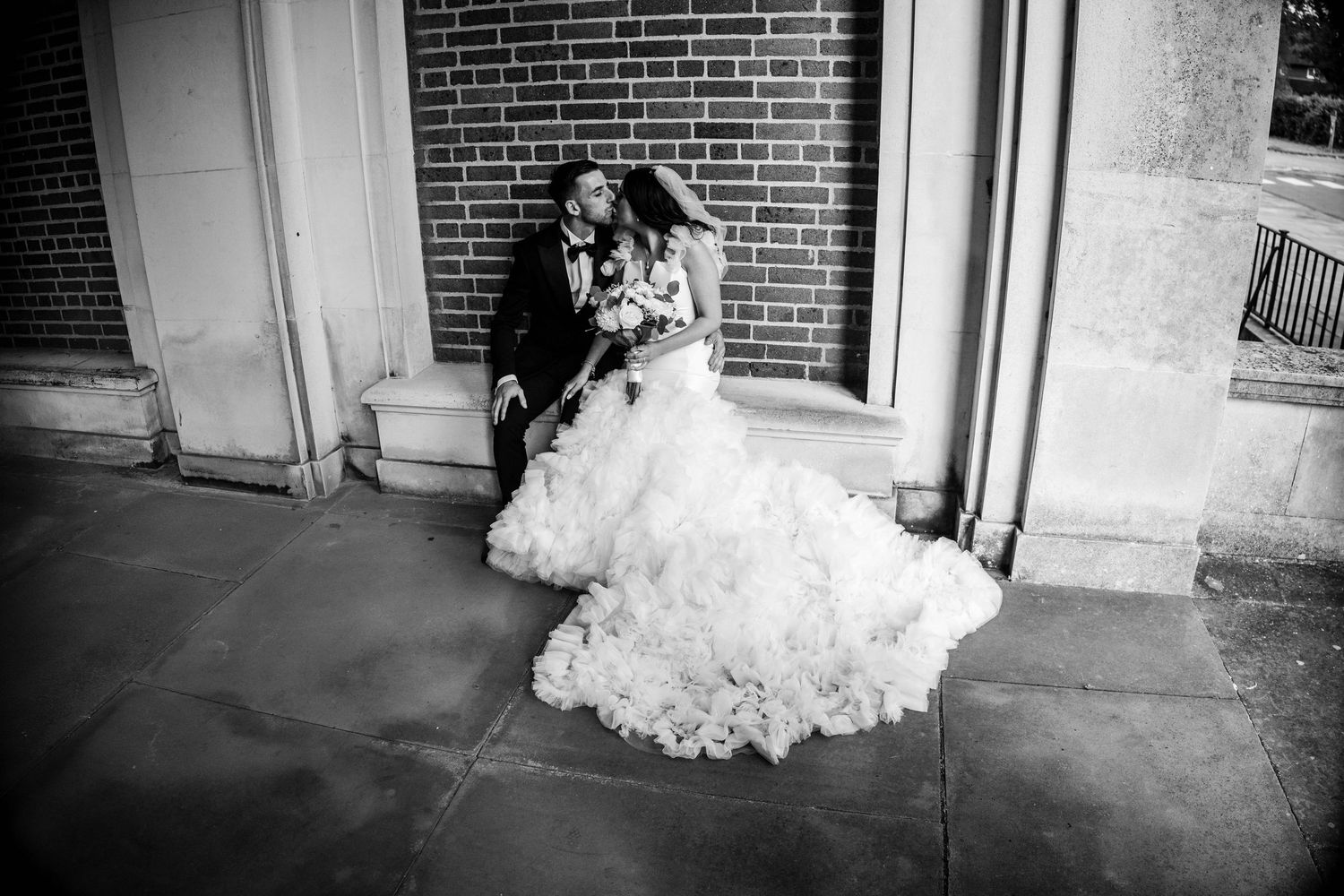 A romantic black and white wedding photo shows a couple embracing on stone steps near a brick wall.