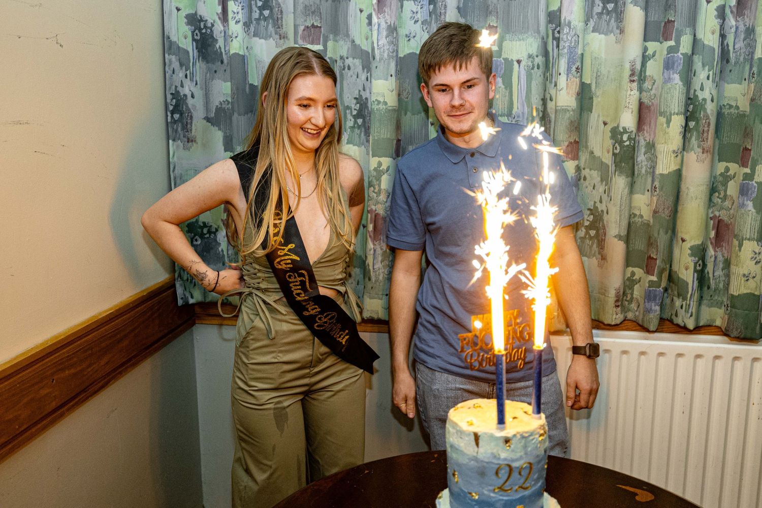 A birthday celebration with sparklers on a cake in front of floral curtains.