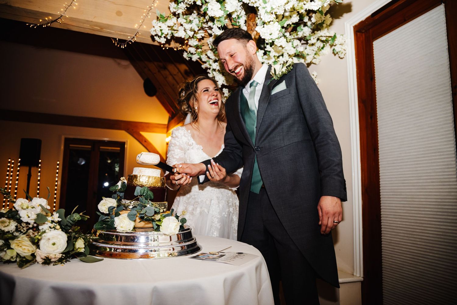 A happy couple shares a laugh while cutting their wedding cake together beneath elegant white floral decorations.