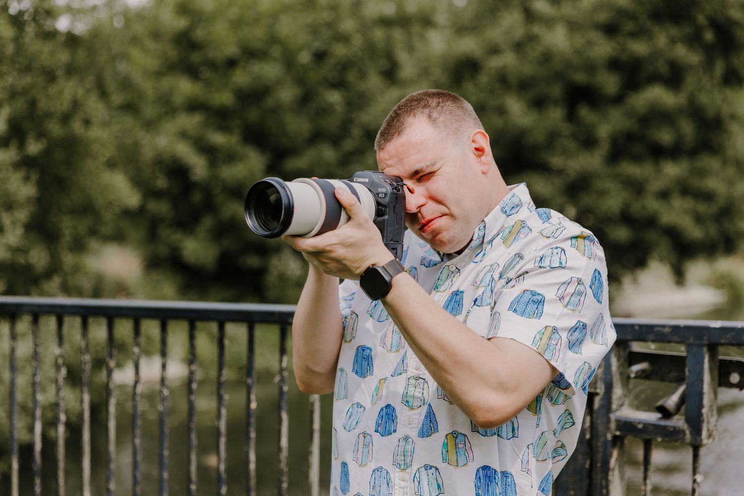Professional photographer in a blue patterned shirt takes outdoor photos with a telephoto lens in a park setting.