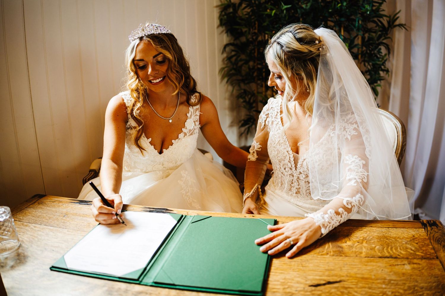 Two people in white wedding dresses sign documents together at a wooden table during a ceremony.