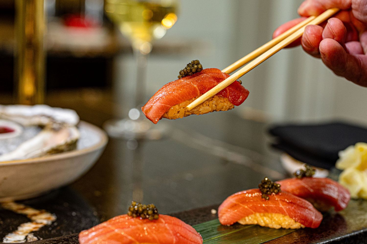 Chopsticks picking up fresh sushi pieces garnished with caviar and sauce against a dark background.