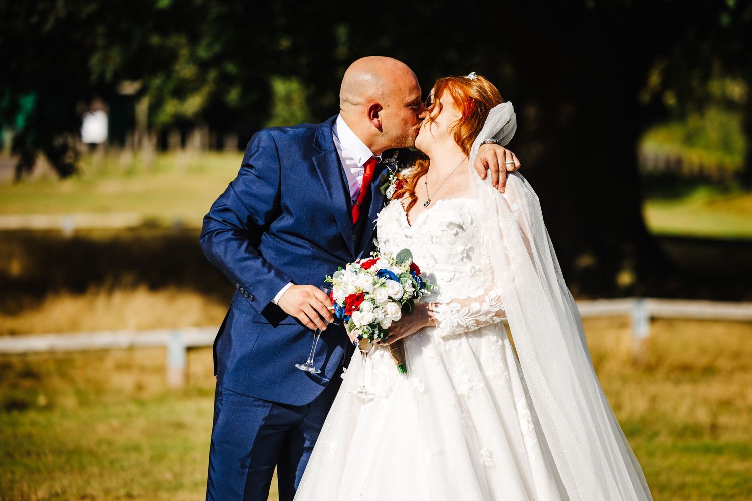 A couple in wedding attire embraces in a sunlit outdoor setting with trees and natural scenery in the background.